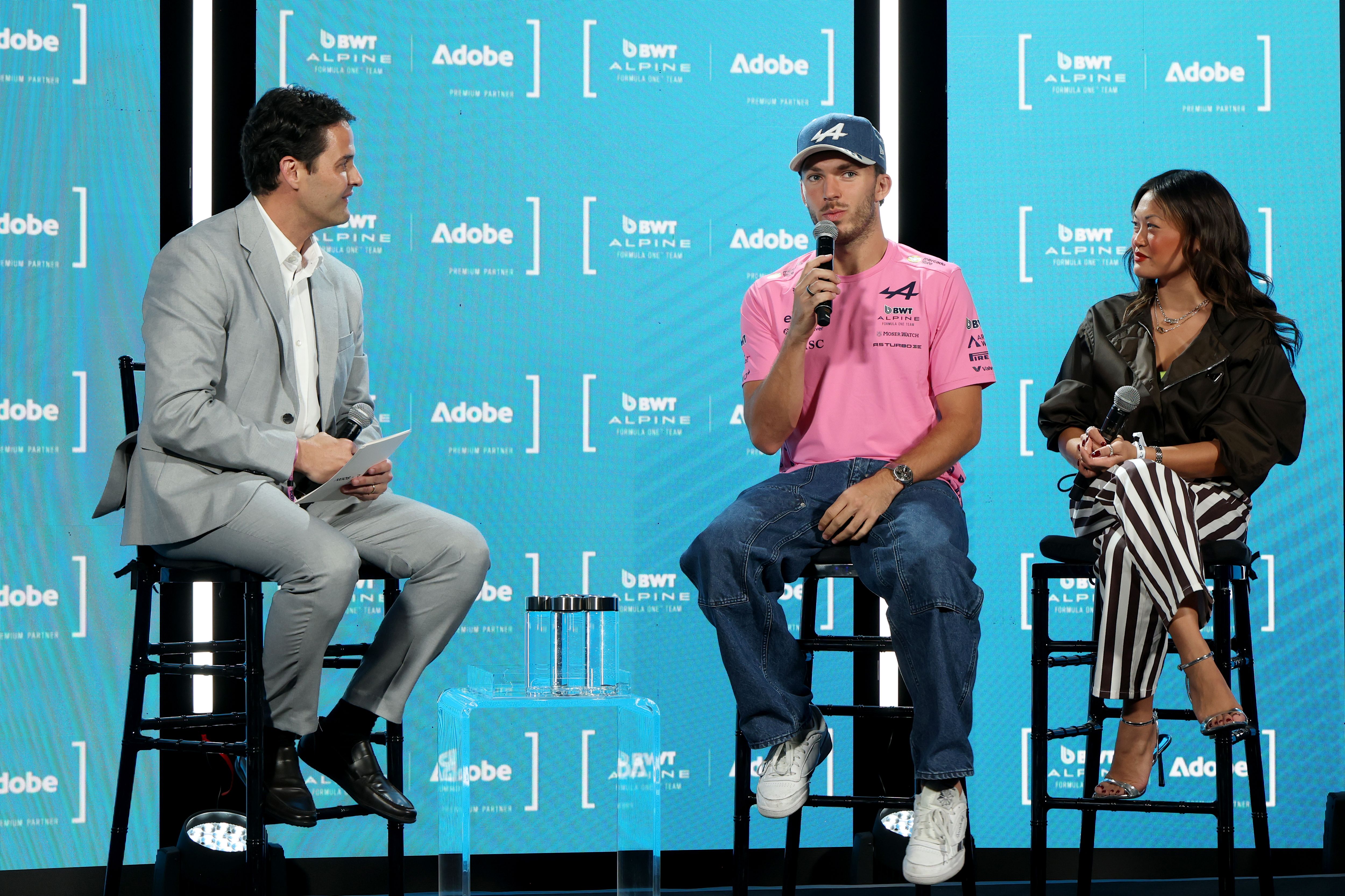 Three-person panel on a bright blue stage with Adobe and Alpine logos. Left: man in a light gray suit; center: man in pink shirt and cap with a mic; right: woman in a black jacket and striped pants.