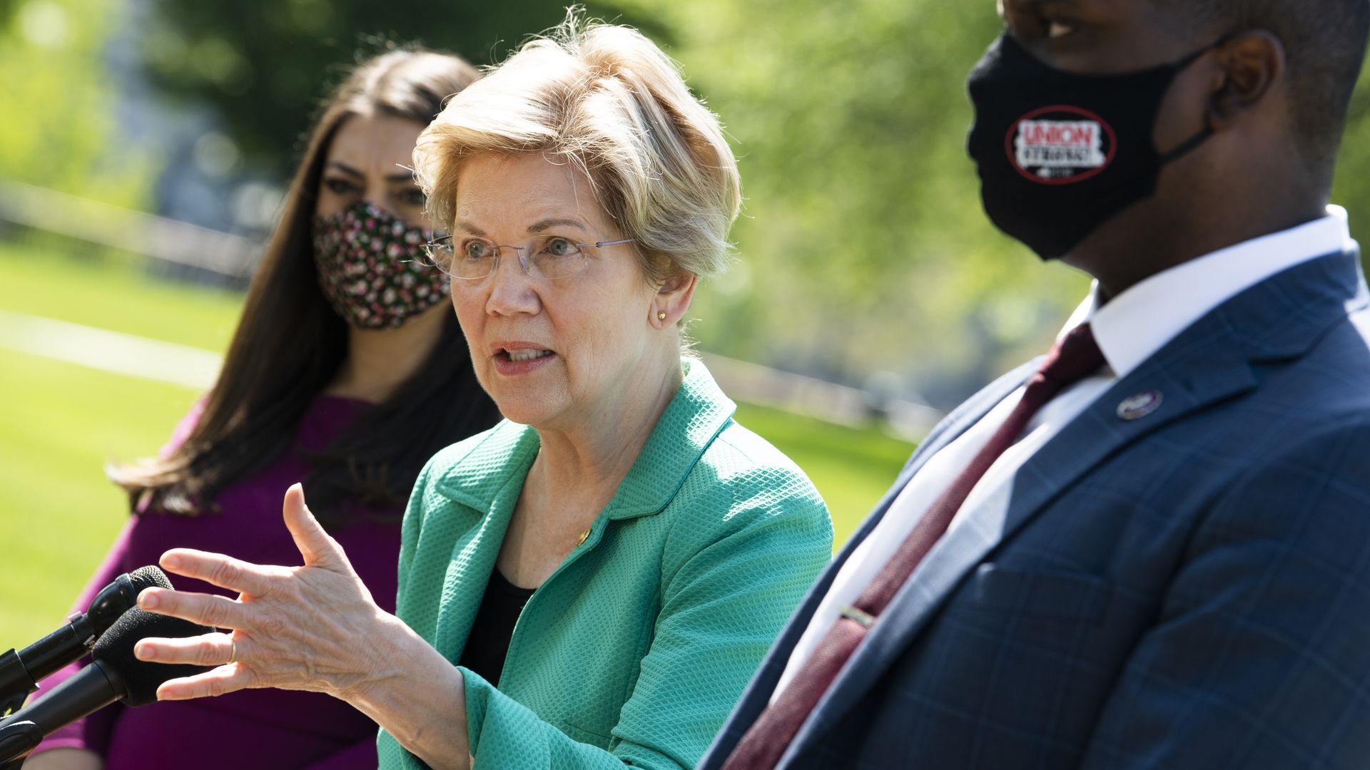 Photo of Elizabeth Warren wearing a green blazer and speaking between two masked representatives