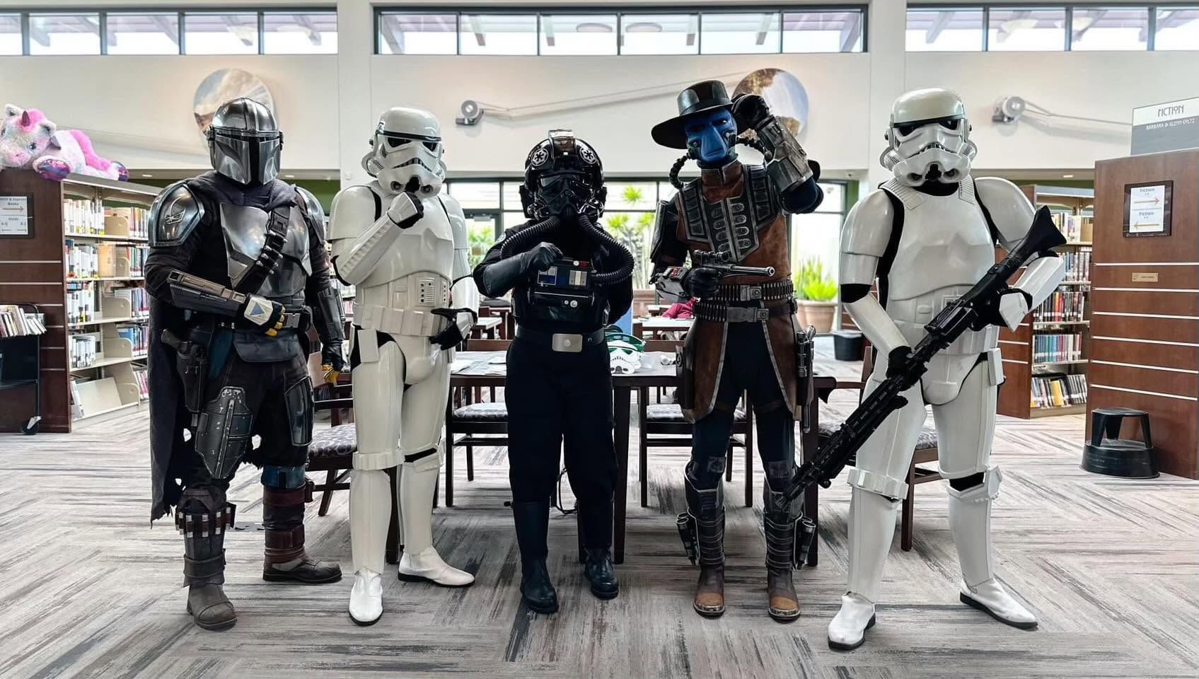 Five Star Wars cosplayers stand in a library: a dark Mandalorian, two white Stormtroopers, a black armored trooper with hoses, and a blue-skinned steampunk fighter with a top hat.