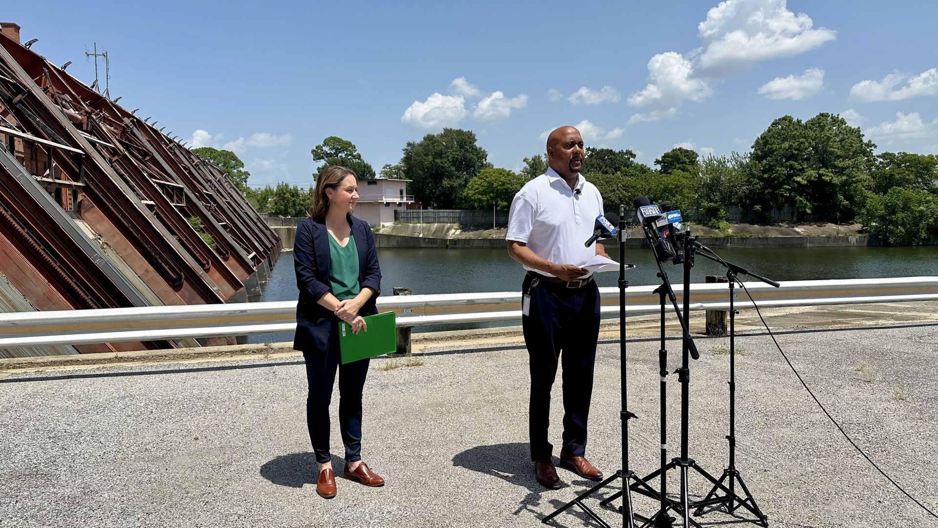 Man in a white polo speaks into multiple microphones while a woman with a green folder stands beside him by a river. A red dam slants on the left; trees and blue sky with clouds fill the background.