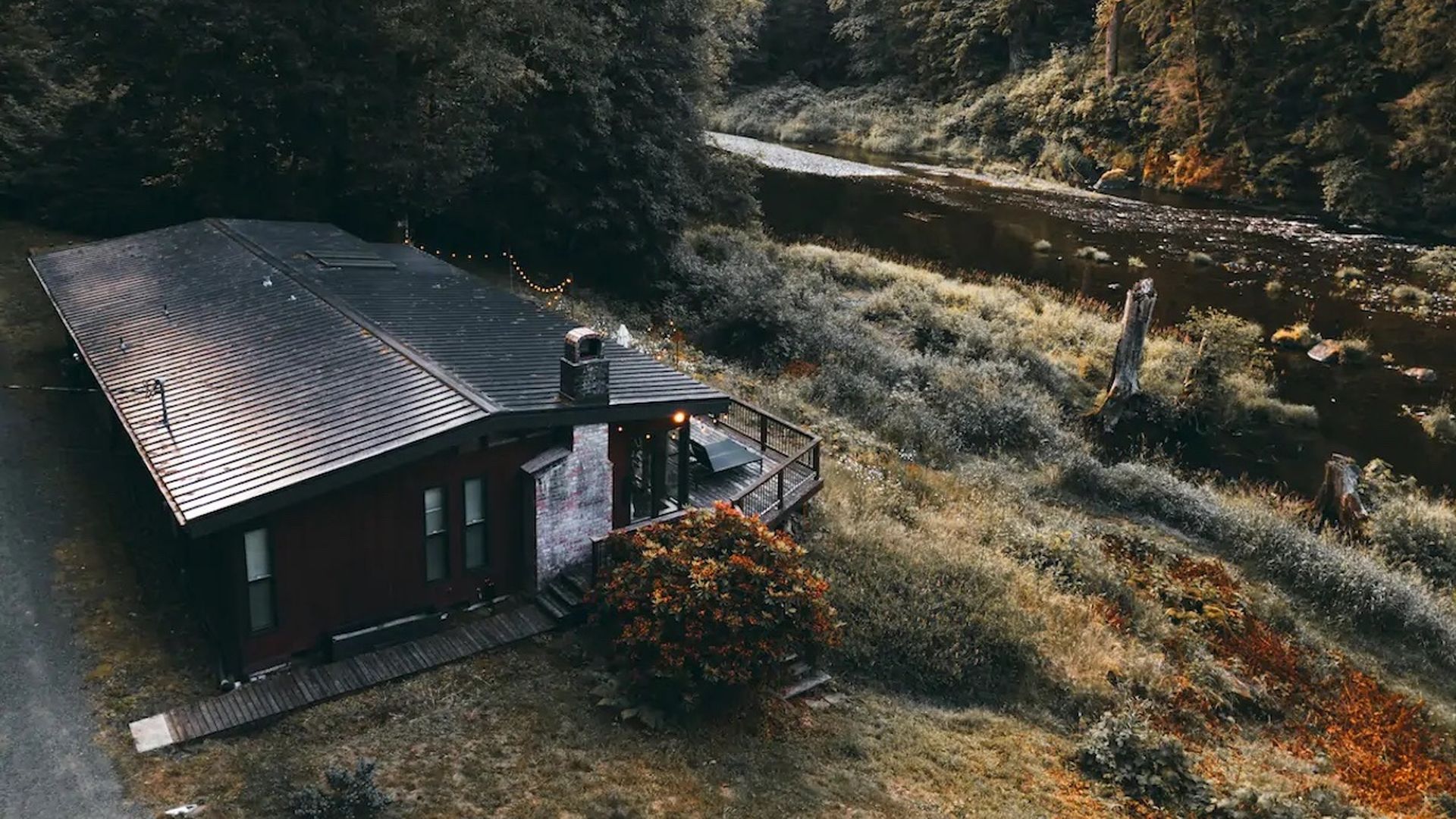 An aerial photo of a cabin with a wrap-around patio alongside a riverfront.