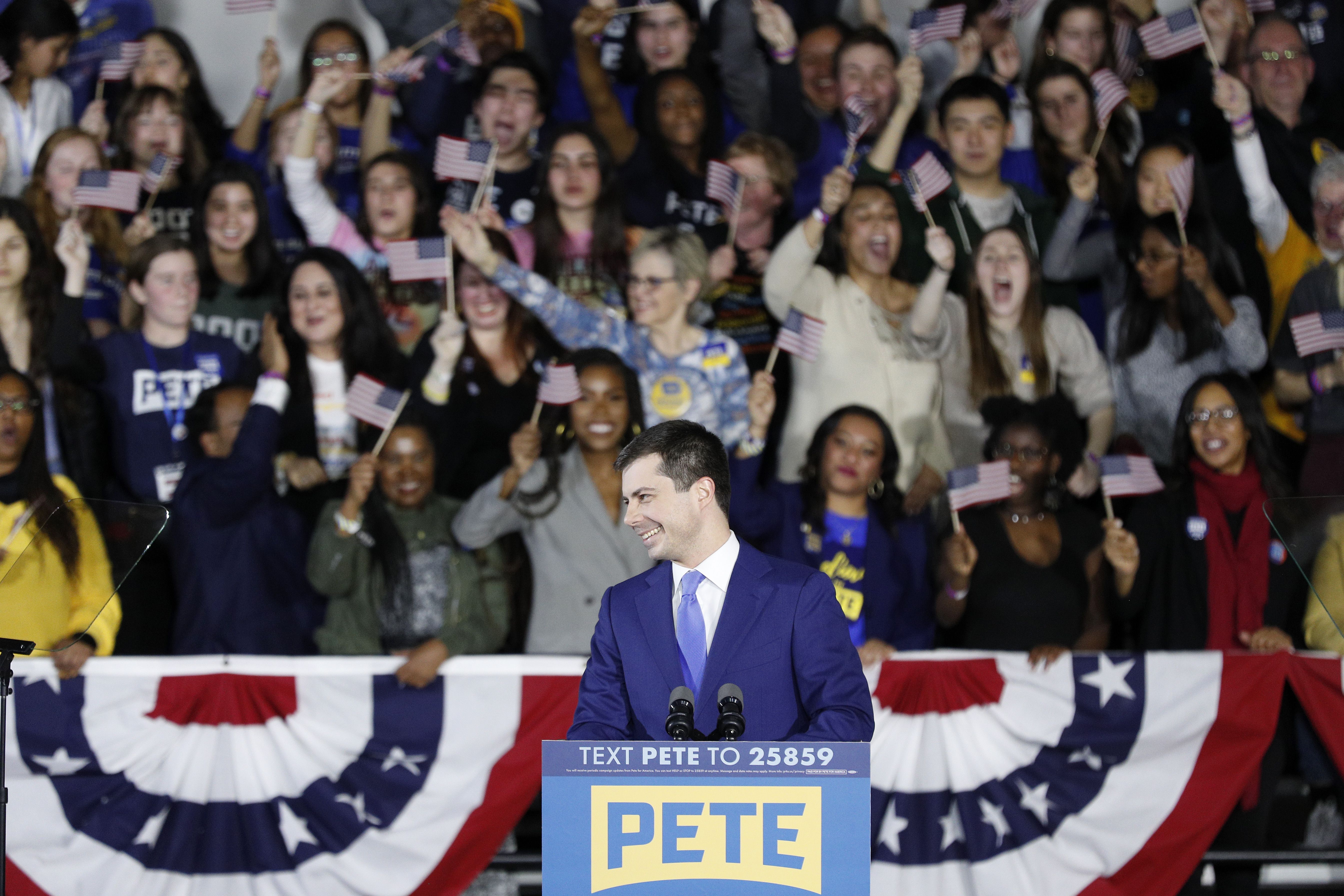 Pete Buttigieg stands at a lectern with a crowd cheering behind him. Several people wave American flags.