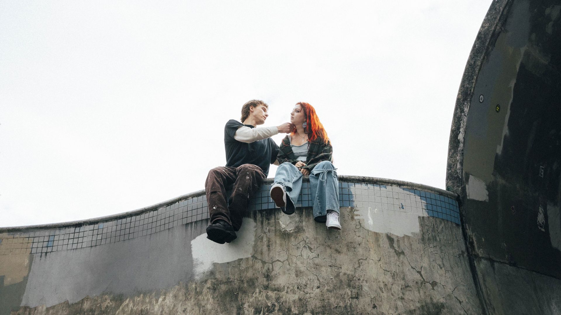 Two young people sit on a cracked, tiled ledge of a crumbling building, facing each other. The man touches the woman’s neck, with a bright sky behind and a dark wall on the right.