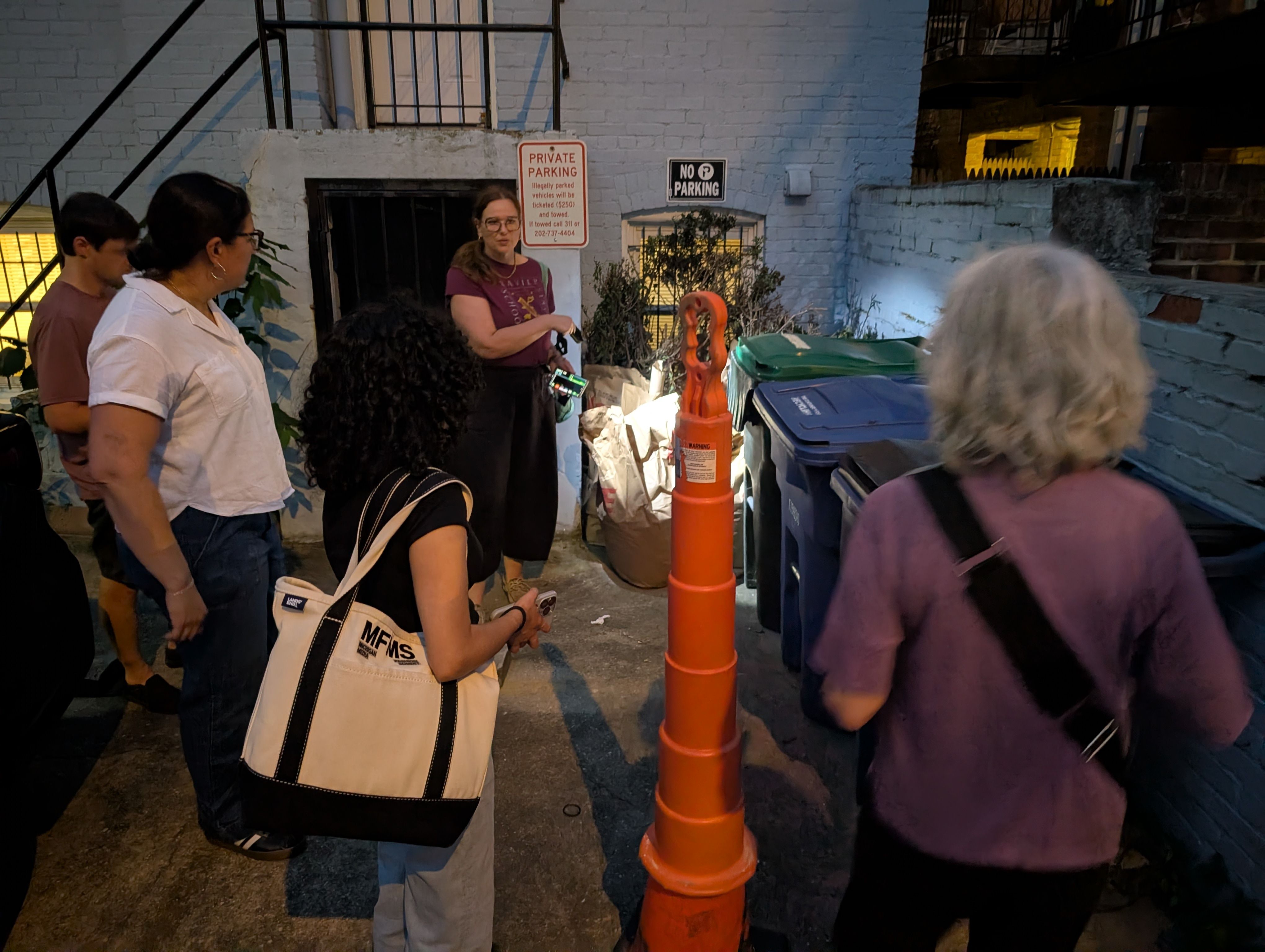 People gather in a narrow alley beside a blue brick wall. An orange cone, trash bins, and signs reading "No Parking" and "Private Parking" are visible as a woman speaks to the group.