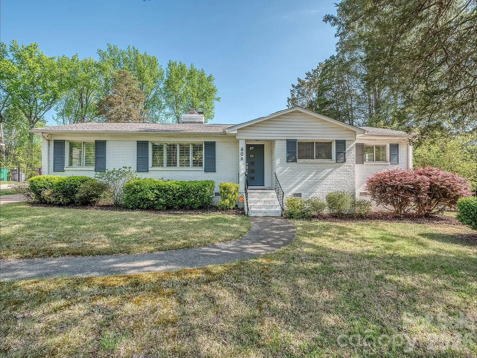 White brick ranch-style house with blue shutters and a dark blue front door, curved walkway, green lawn, trimmed hedges, pink shrubs on the right, and tall trees against a clear blue sky.