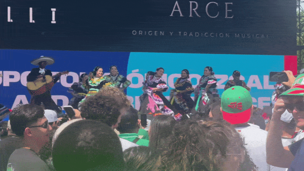 Outdoor stage with a line of dancers in bright traditional outfits performing, a blue banner backdrop, and a crowded audience filming with phones in front of the stage.