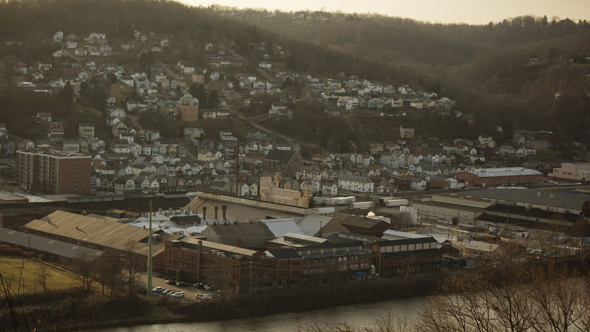 Industrial buildings by a river with a hillside neighborhood of houses and trees in muted evening light