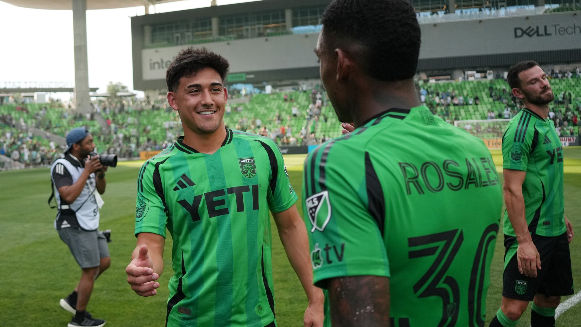 Three soccer players in bright green striped jerseys with the YETI sponsor stand on a grass field in a stadium. One smiles at the camera while two face away; a photographer and green seating fill the background.