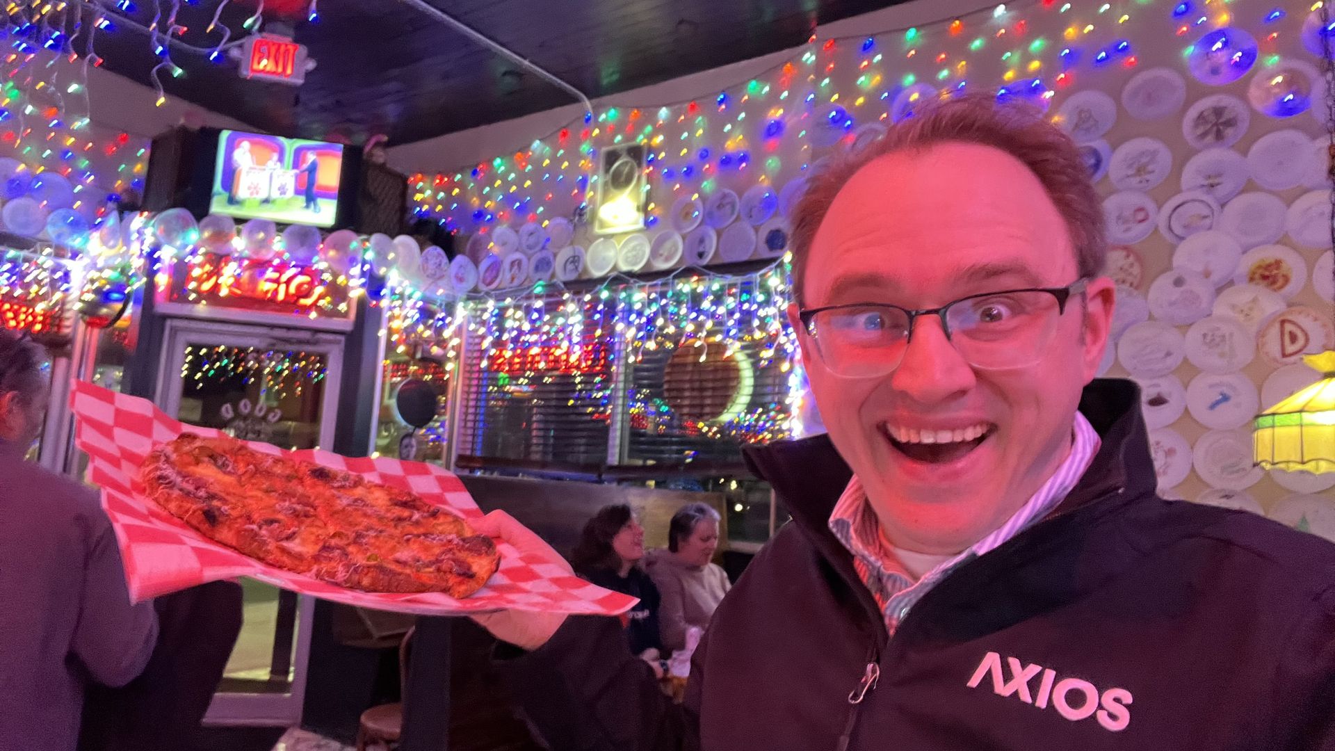 Man wearing glasses and an Axios jacket excitedly holding a pizza on red checkered paper in a brightly decorated restaurant with colorful string lights and plates on the walls.