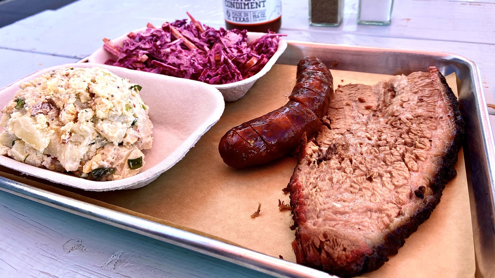 A metal tray of food that includes potato salad, purple slaw, a scorched sausage and a slab of moist brisket.