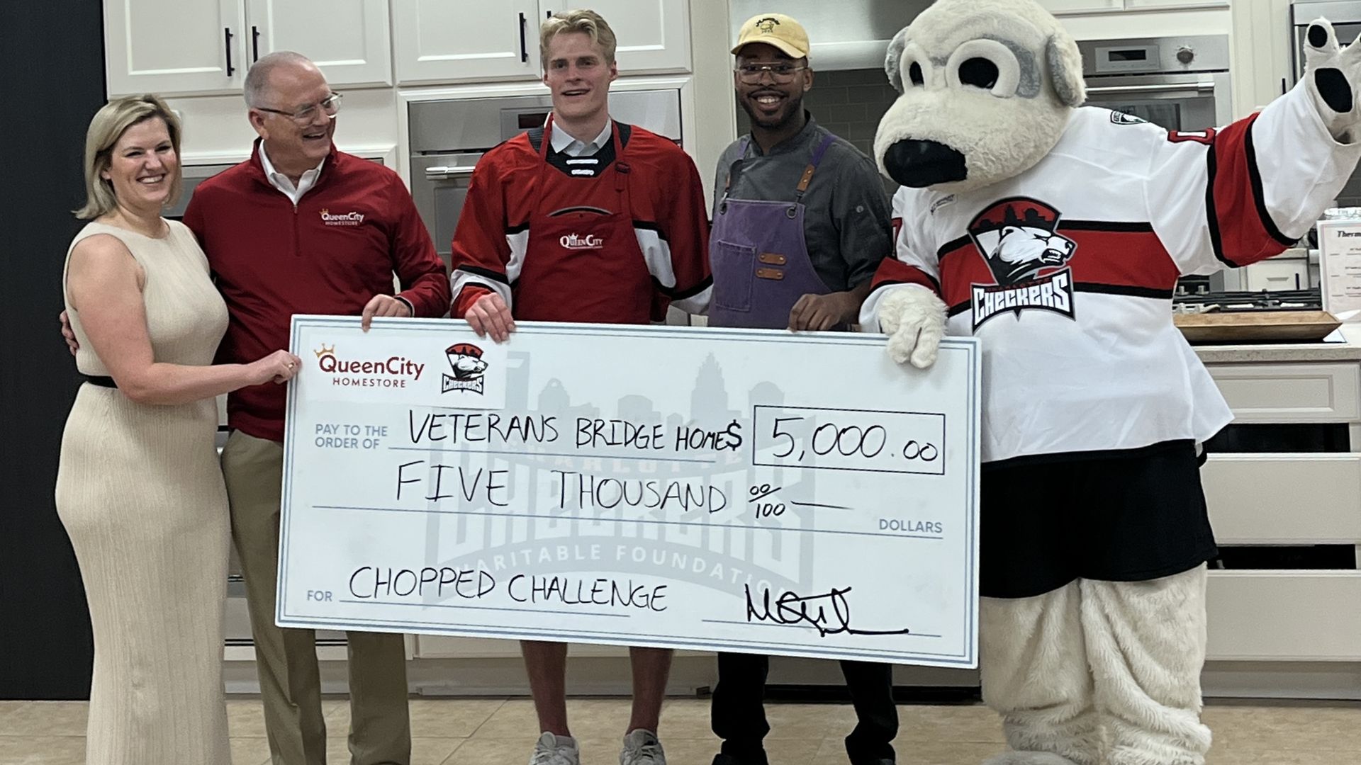 Five people and a large mascot pose in a bright white kitchen, holding a giant check for Veterans Bridge Homes for $5,000 for the "Chopped Challenge" featuring Queen City and Checkers logos.