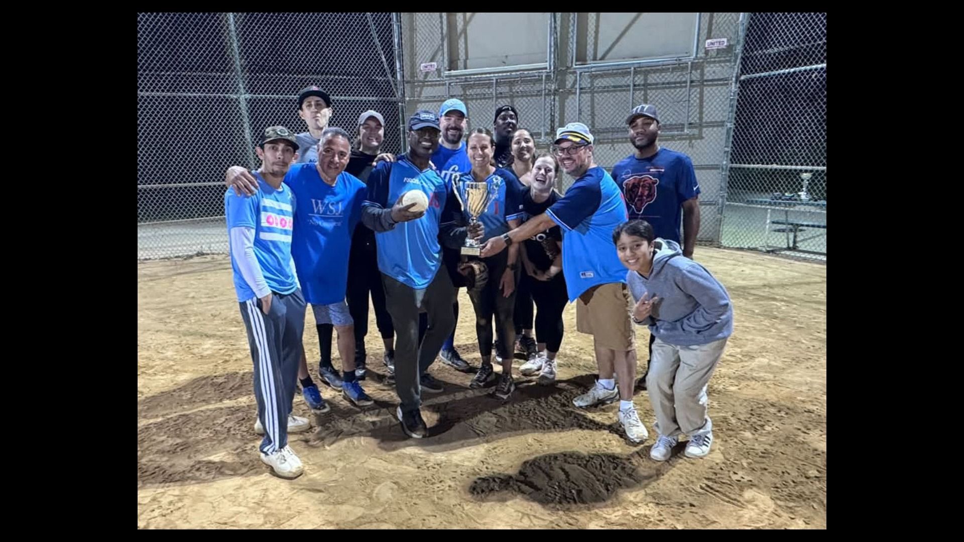 A diverse group of people in blue jerseys stand on a dirt baseball field, posing with a large trophy and a ball, smiling for the camera in front of a chain-link fence.