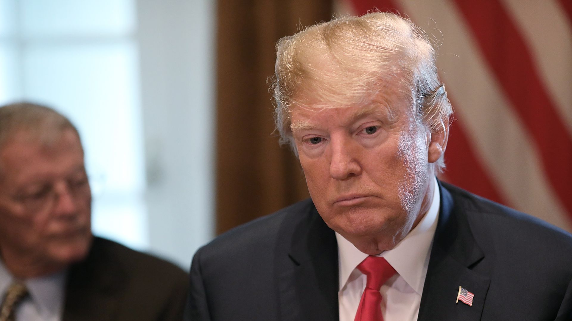 Donald Trump looks down before an American flag while sitting.