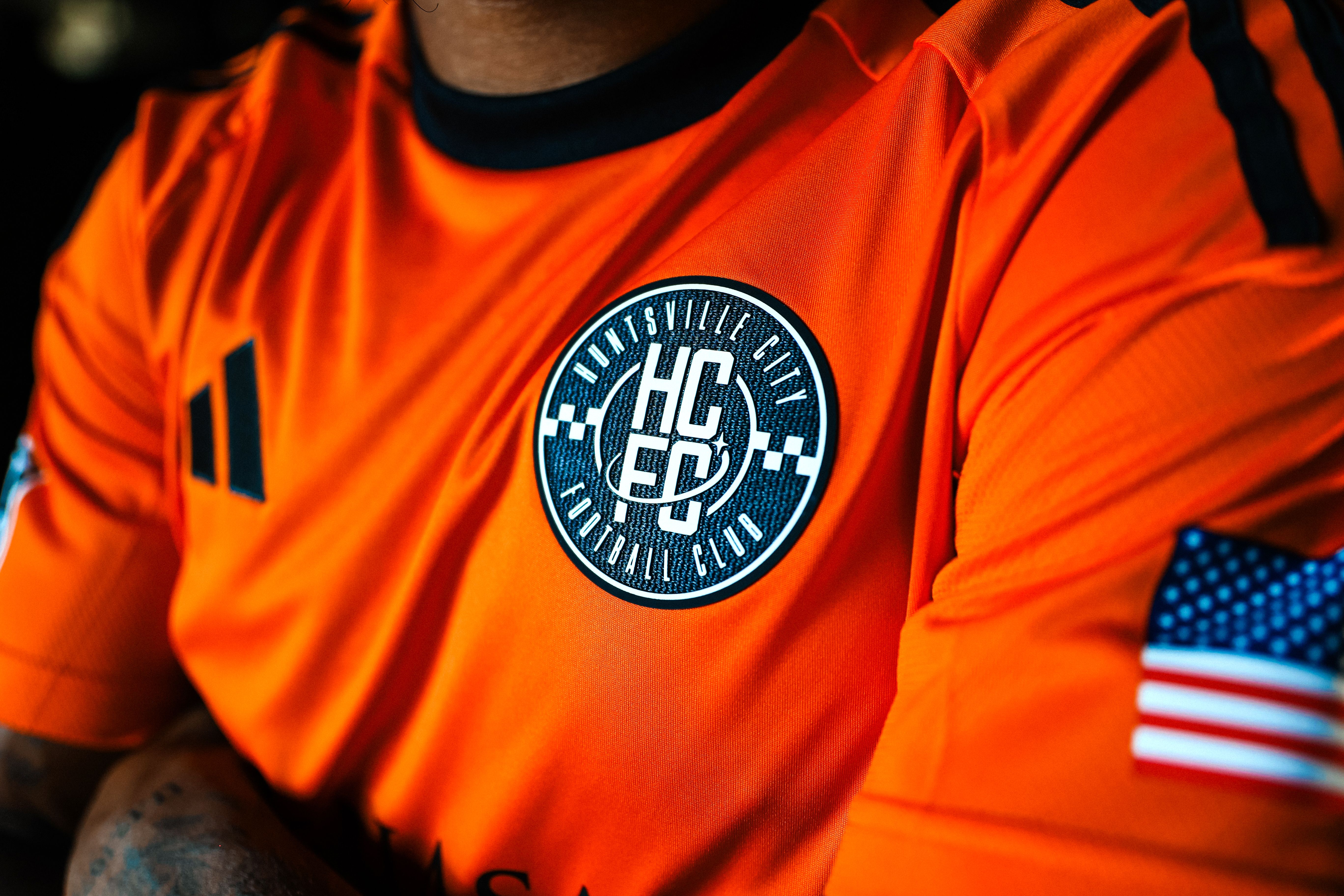 Close-up of an orange Huntsville City Football Club jersey with black stripes, club logo in white and black on chest, Adidas logo, and an American flag patch on the sleeve.