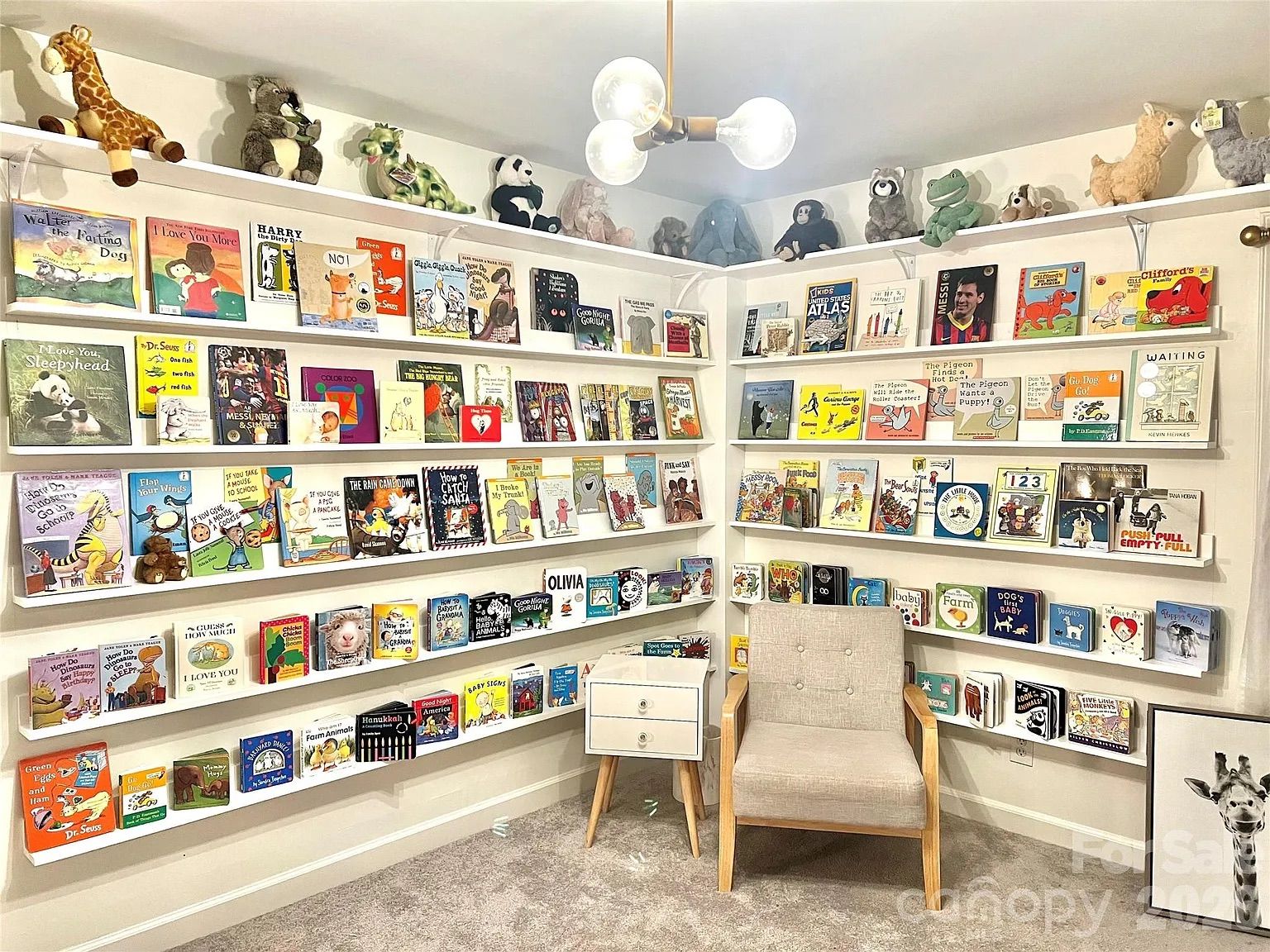Children's reading nook with white shelves filled with colorful books, stuffed animals on top shelves, a beige armchair, and a white side table under a modern ceiling light.