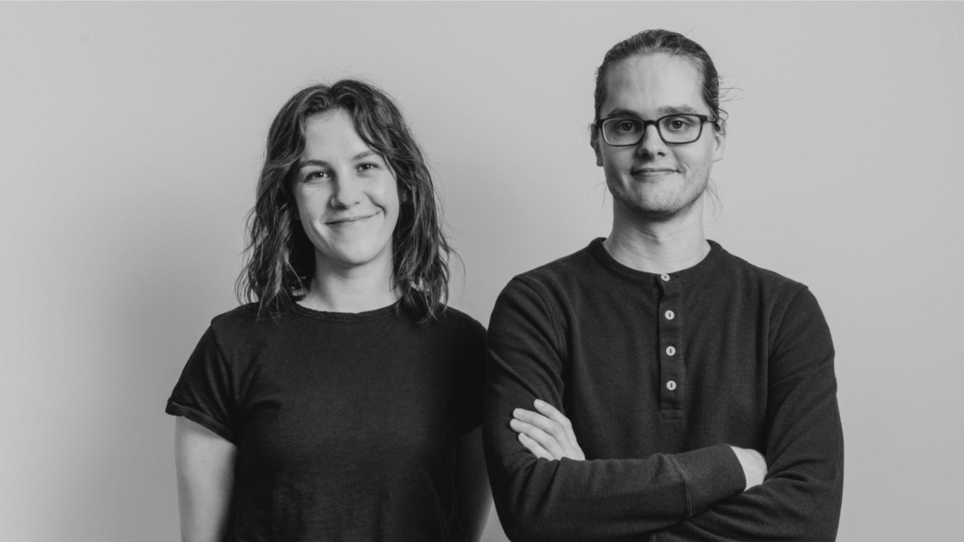 Two people standing side by side against a gray background: a smiling woman on the left in a black T-shirt and a man with glasses on the right in a dark henley, arms crossed.
