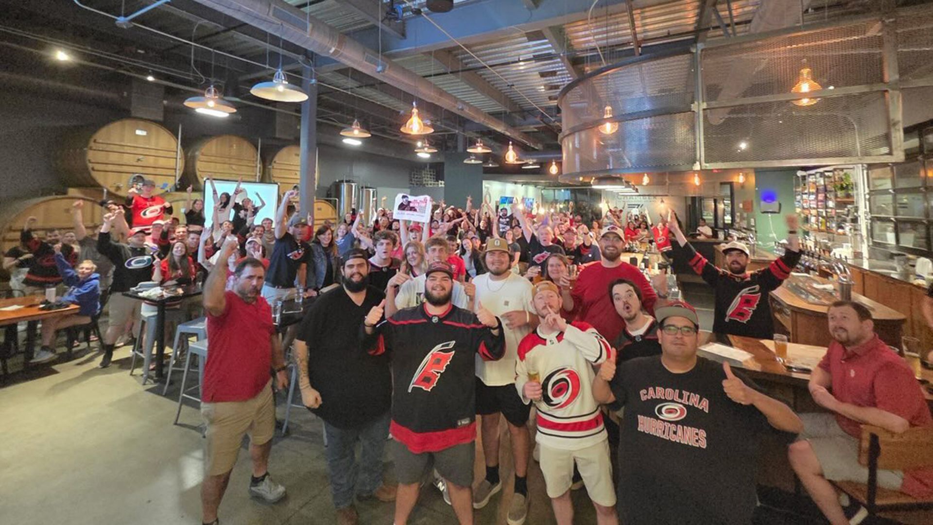 Wide group photo inside a brewery-style venue with exposed ceiling, hanging lights, and large wooden barrels. People cheer and pose, many in Carolina Hurricanes jerseys.
