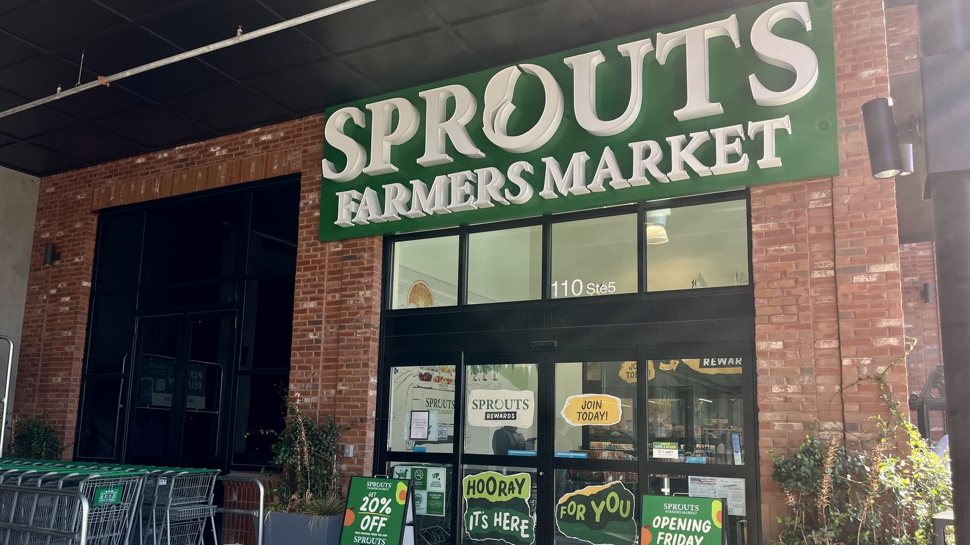 Entrance of Sprouts Farmers Market with green and white signage, shopping carts on the left, and promotional signs for 20% off and an opening on Friday, Aug. 22nd in front of glass doors.