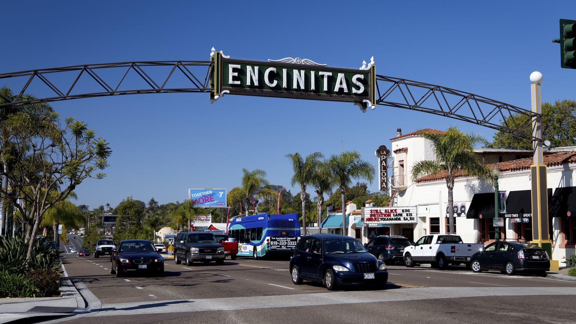 Street view of Encinitas with a large arched sign reading "ENCINITAS" over the road, cars driving, palm trees, and buildings including La Paloma theater under clear blue sky.