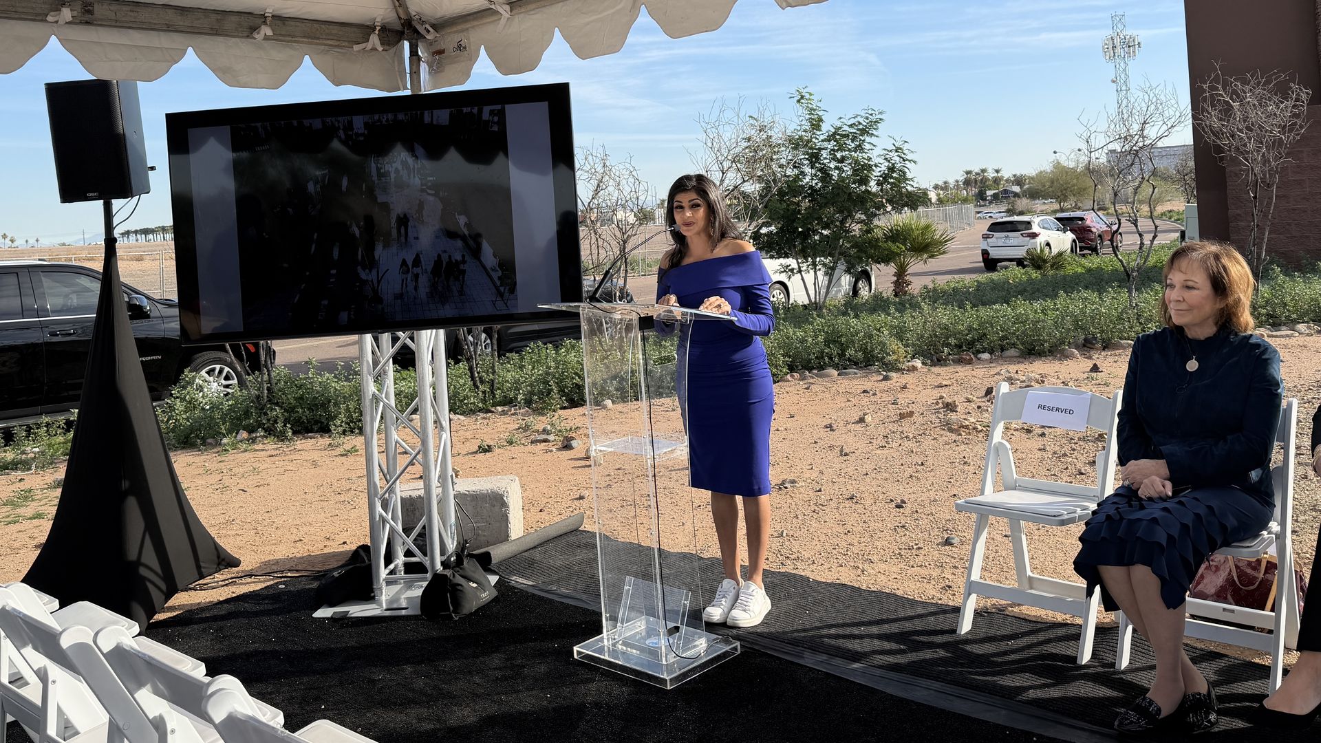Woman in a blue dress and white shoes speaking at a clear podium under a tent, with a large screen and a seated woman in a dark blue outfit nearby, in a sunny outdoor setting.