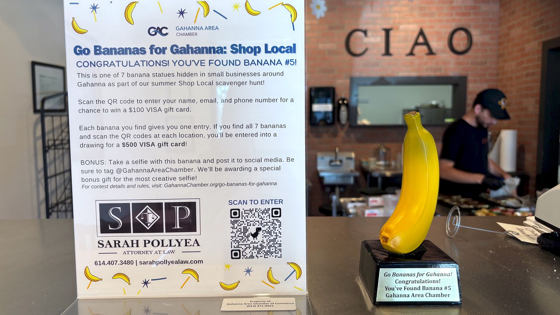 Sign and yellow banana statue on black base on counter promoting Gahanna Area Chamber's "Go Bananas for Gahanna" Shop Local scavenger hunt with store interior and worker in background.