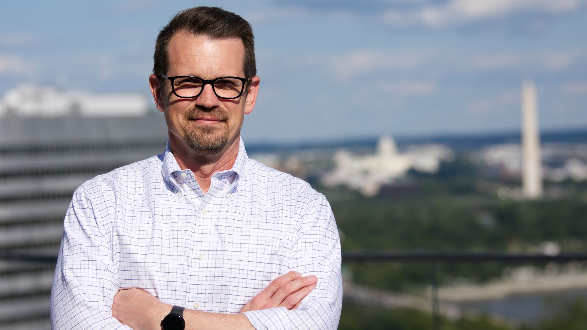 Jason Brown, a man, stands on a balcony overlooking Washington, D.C. He's in a white dress shirt. His arms are crossed. He is wearing glasses.