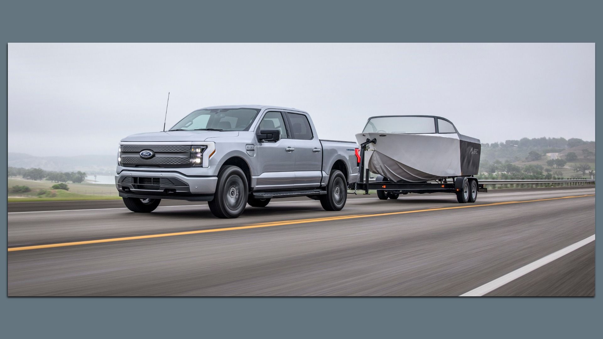 Silver Ford F-150 Lightning electric pickup truck towing a boat on a highway with overcast sky surrounded by green landscape and trees.
