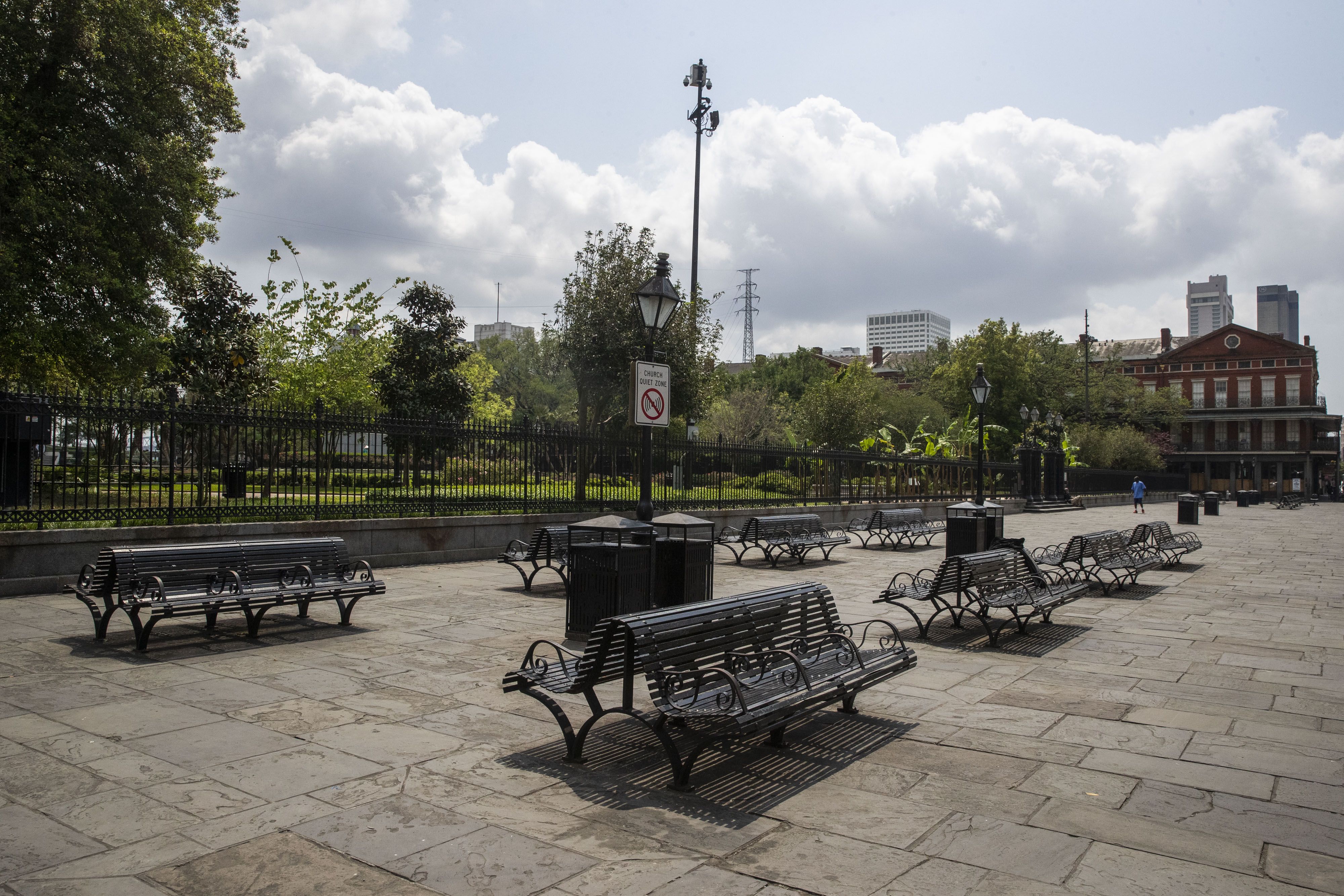 Empty black metal benches on a stone-paved plaza under a cloudy sky, with a lamp post and a "Church Quiet Zone" sign near a fenced green park and buildings in the background.