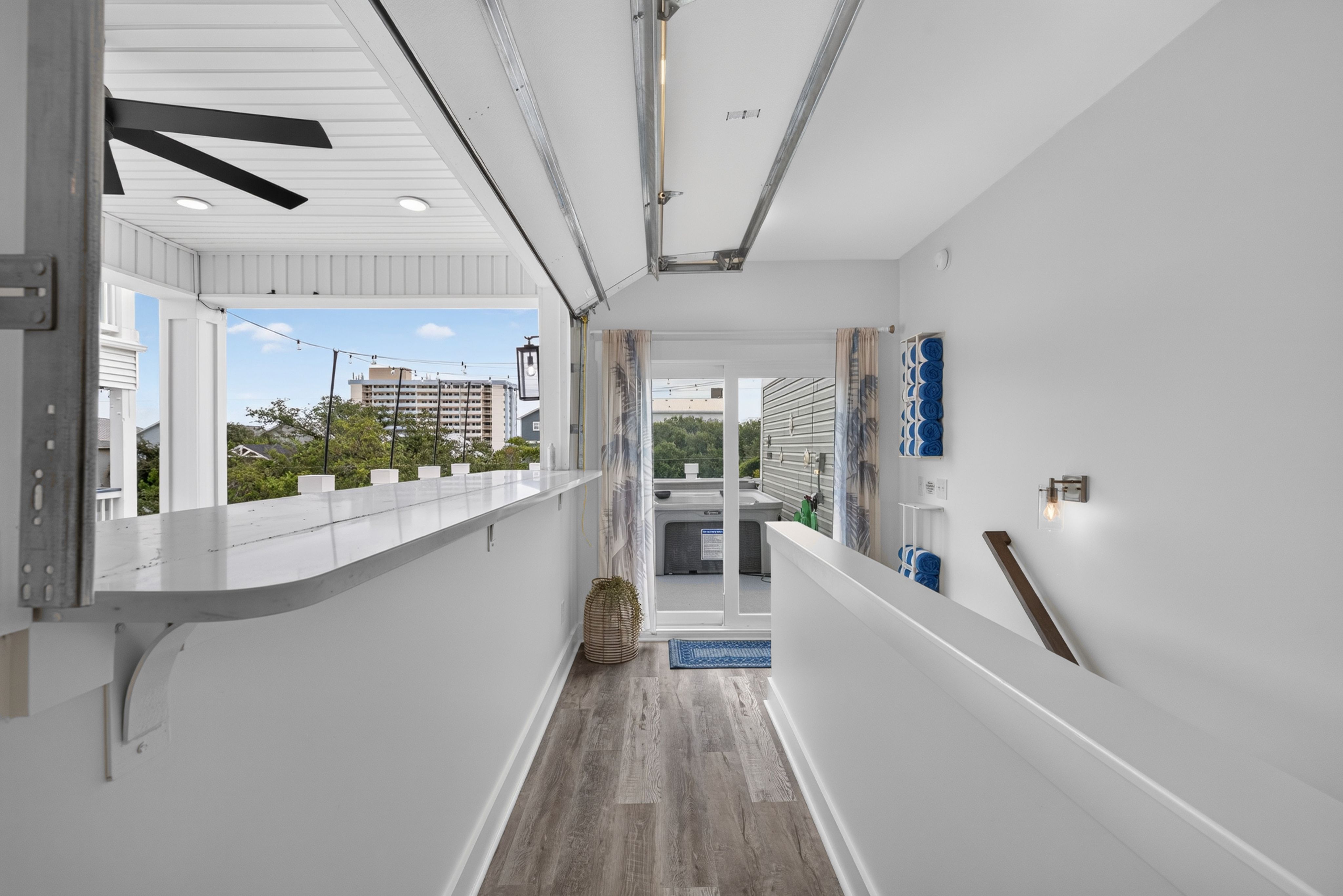 Bright laundry/utility space with white walls and ceiling, white counter on the left, washer/dryer at the far end, open doorway to blue sky, black ceiling fan blades, and blue towels on a wall rack.