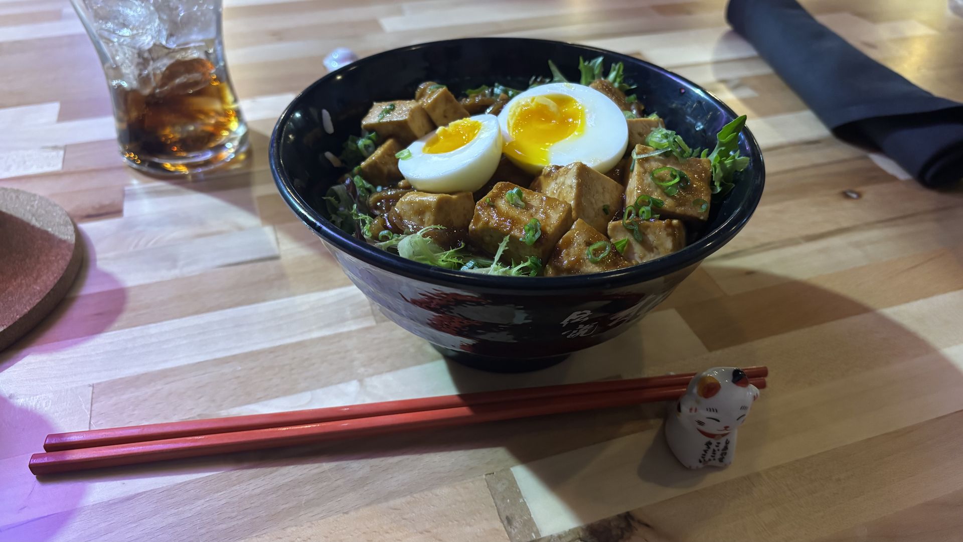 Black bowl with tofu, greens, noodles, scallions, and a halved soft-boiled egg on a light wood table with red chopsticks resting on a cat-shaped chopstick holder and a glass of iced soda nearby.