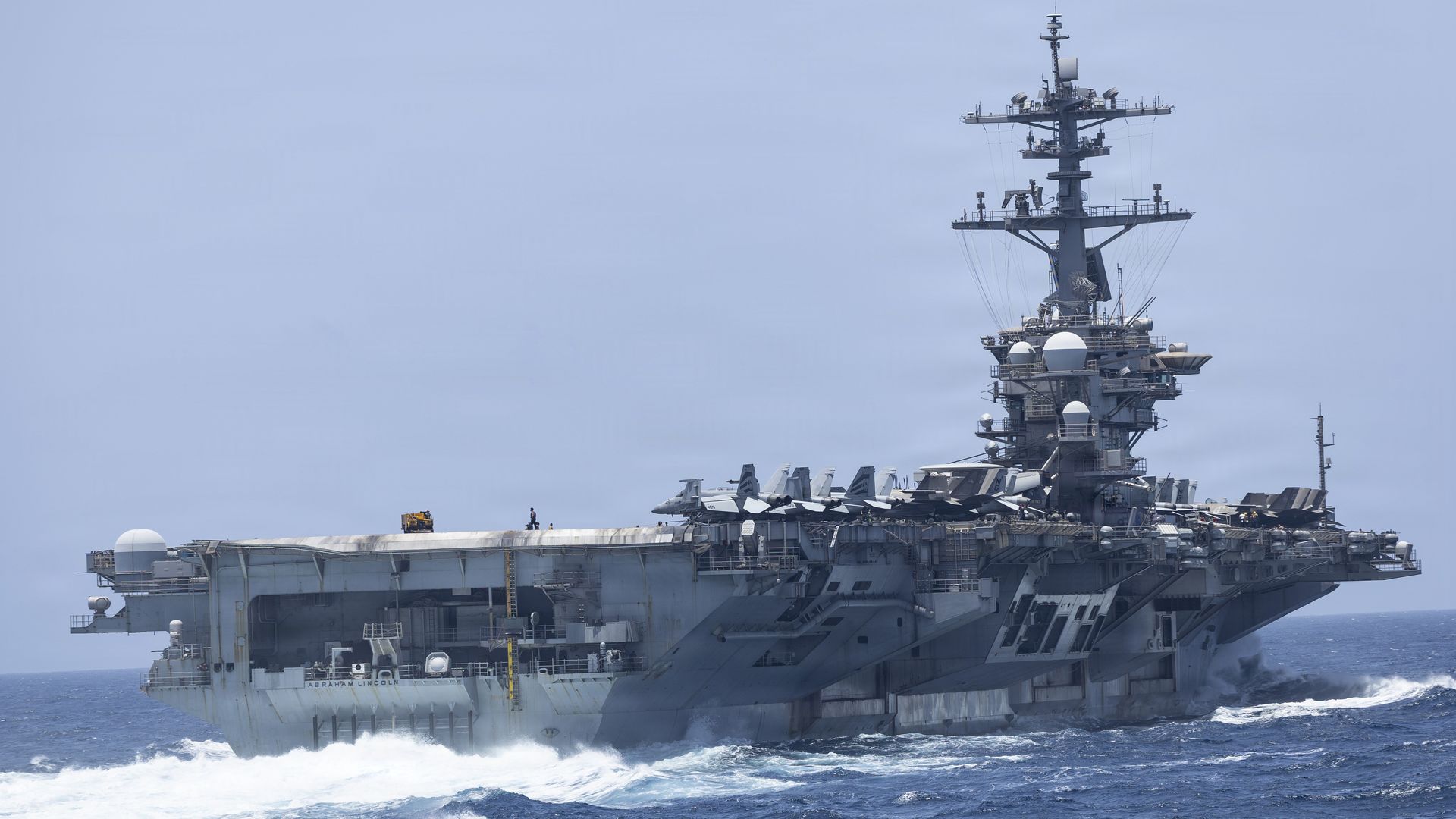 Side view of a large gray aircraft carrier at sea, deck lined with fighter jets, a tall island with antennas, blue sky above; hull marked with the ship name Abraham Lincoln.