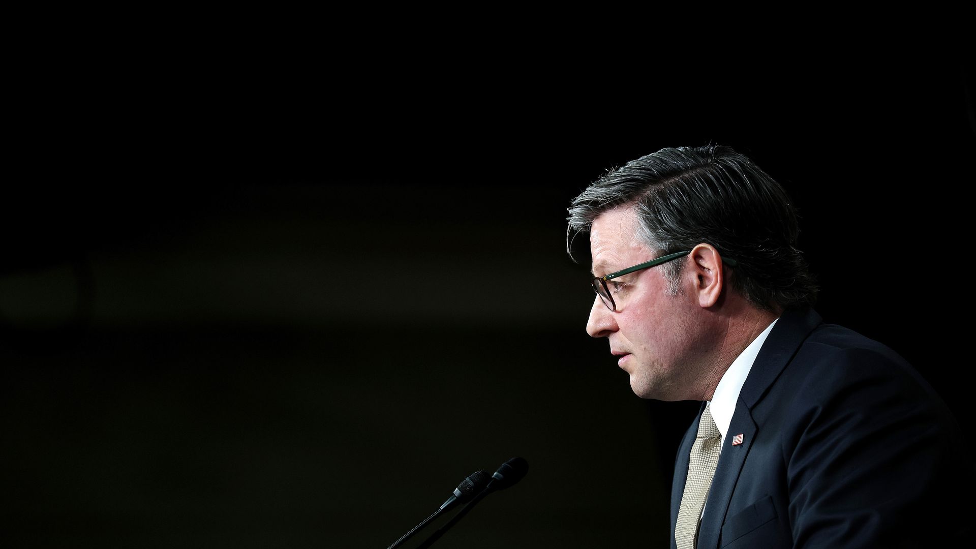 Partial side profile of a man in a dark suit and tan tie, wearing green-framed glasses, speaking at a podium with two microphones against a black background.