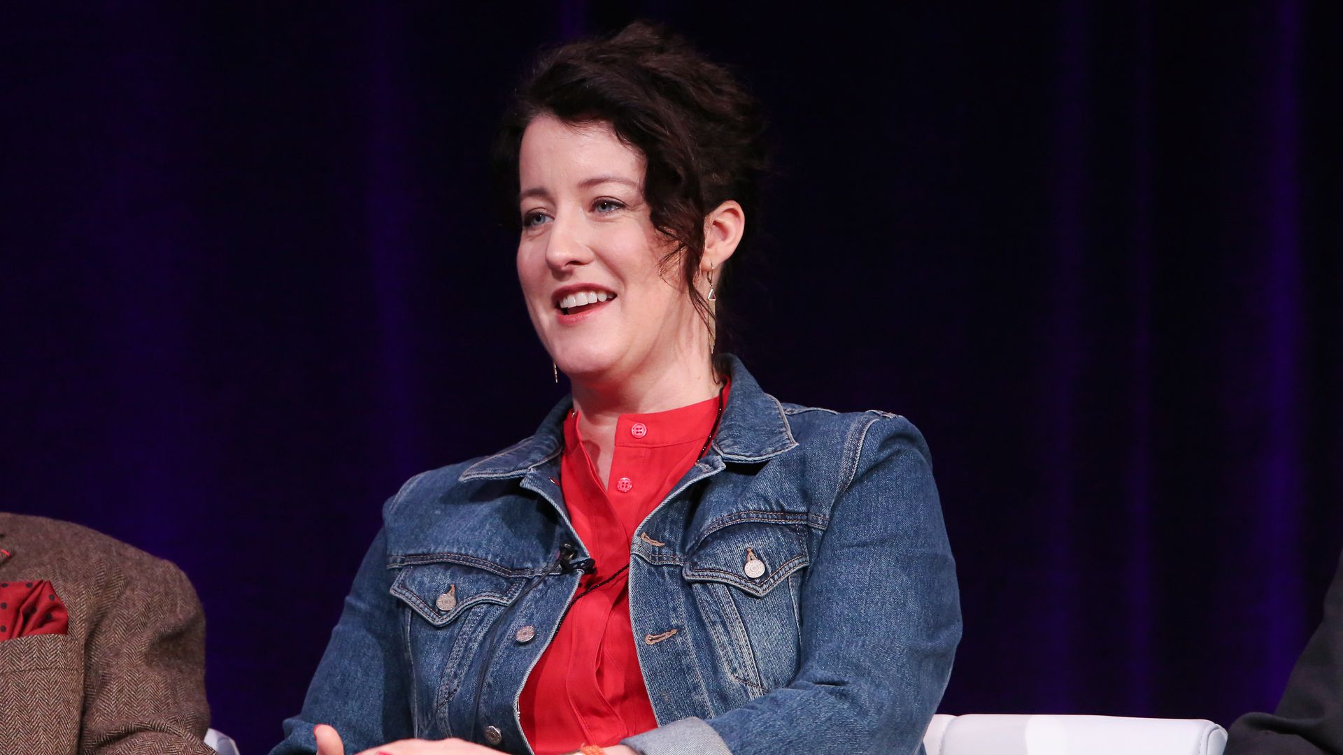 A photo of a person wearing a red shirt and jean jacket speaking on a panel.