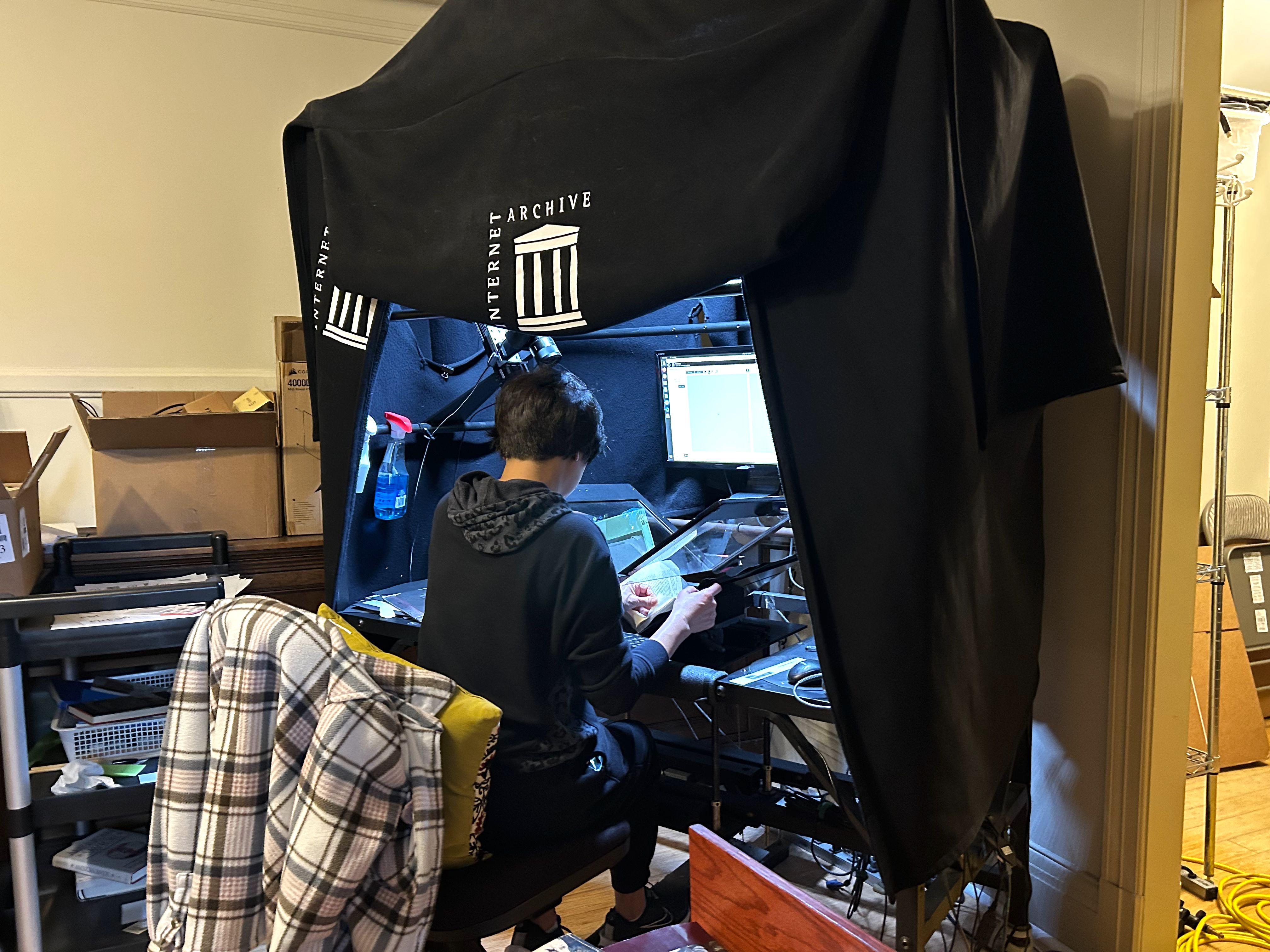Person sitting in a black hooded sweatshirt at a scanning station with a black cloth labeled "Internet Archive," digitizing pages under bright light with monitor and camera setup.