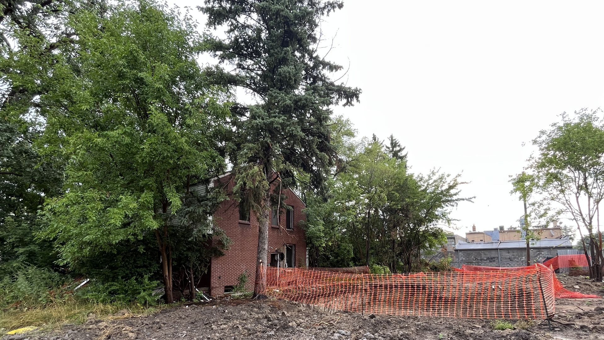 A home is shown hidden among trees, with a caution fence around its yard. Lots of dirt in the foreground.