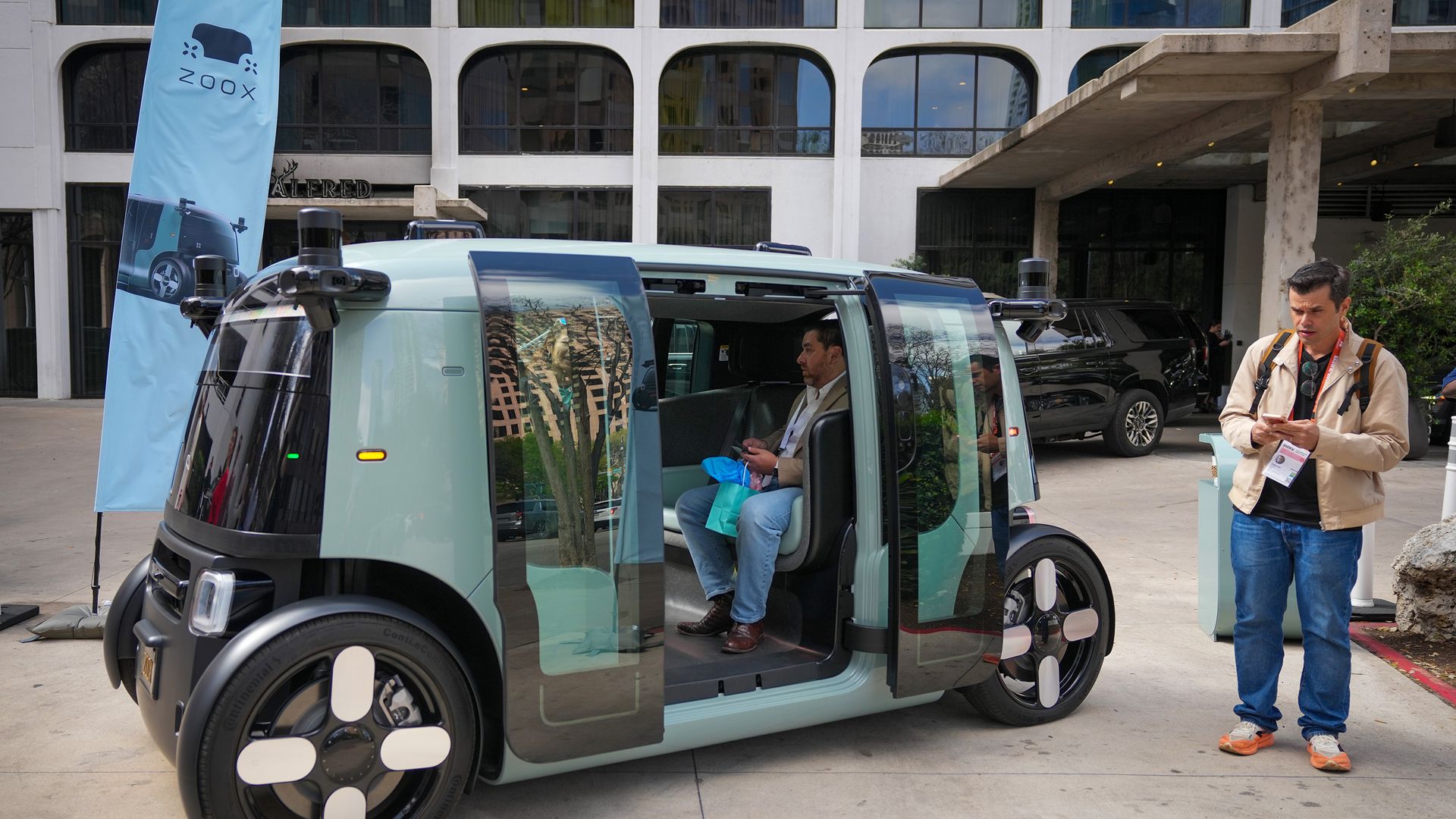 Light blue autonomous vehicle with open doors; a man sits inside holding a bag while another man in a beige jacket stands outside with a badge. A blue Zoox banner and modern building in background.