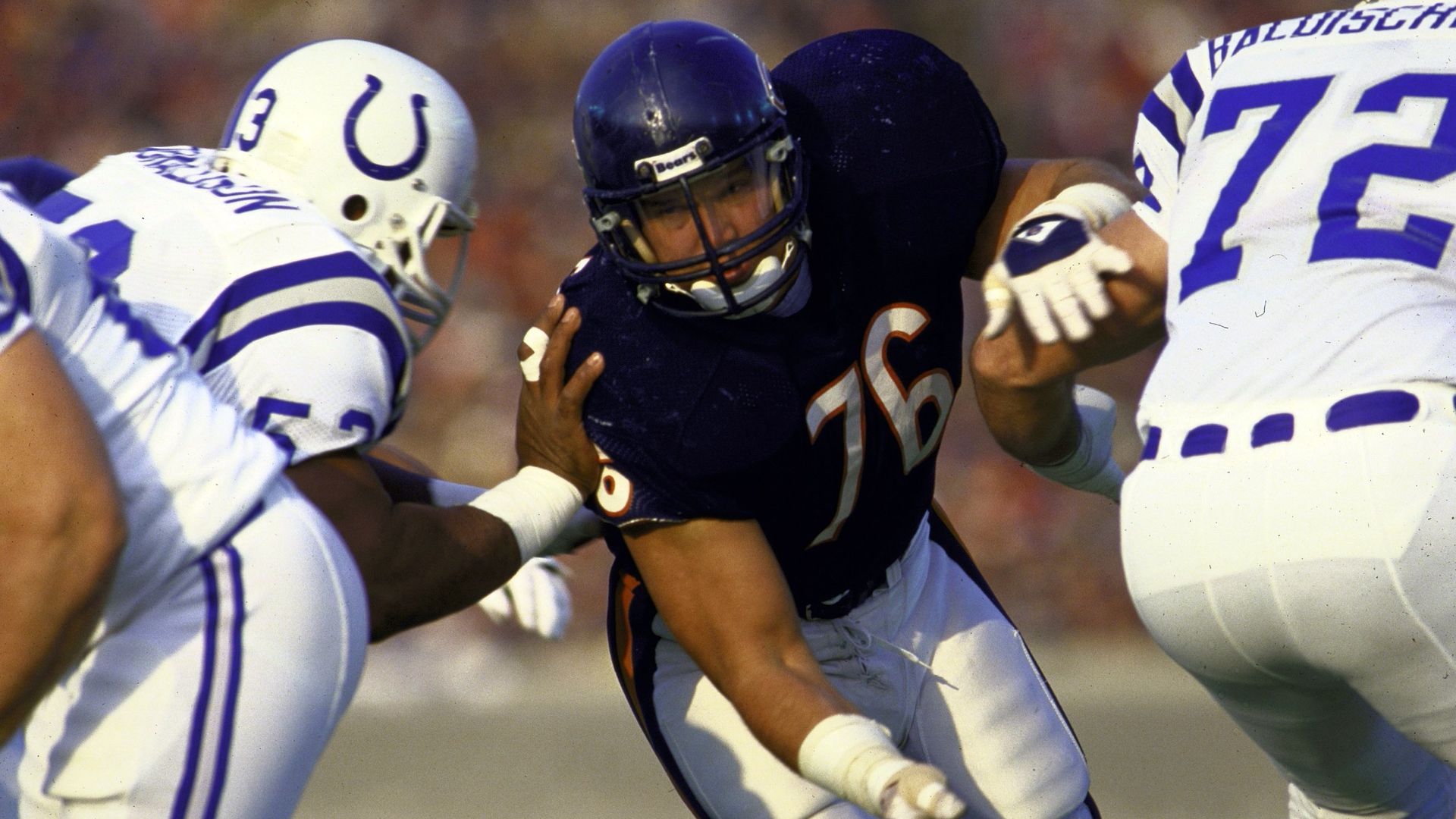 A football player in a dark blue jersey with number 76 pushes against two players in white jerseys with blue horseshoe logos, one with number 53 and the other 72, during a game.