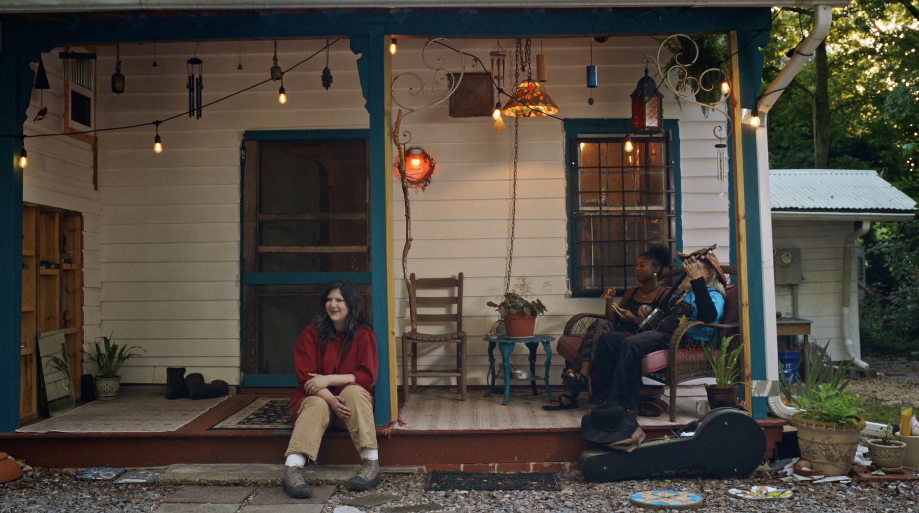 On a white wooden porch with teal trim, a woman in red sits on the steps left while two people on wicker chairs to the right play a guitar. Warm string lights and a colorful lampshade hang overhead.