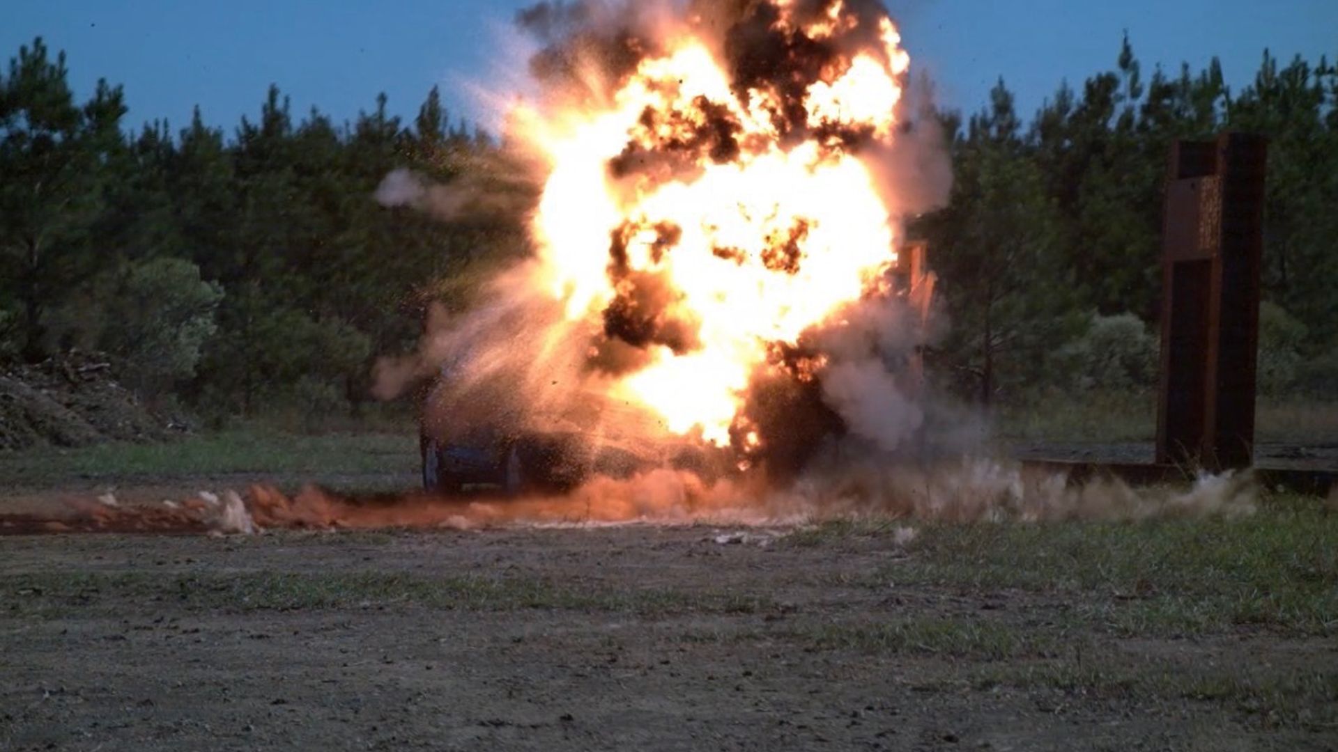 Large fiery explosion with smoke on a dirt ground in front of dense green trees under a clear blue sky, with AEON and DMPG logos in corners.