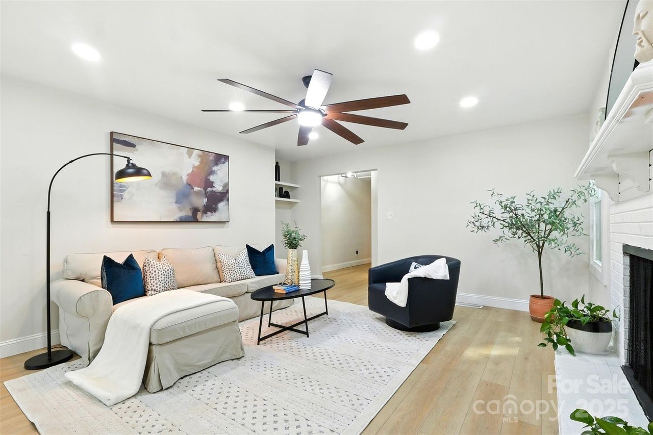 Bright living room with beige sectional sofa, blue and patterned pillows, black swivel chair, black arc floor lamp, abstract wall art, large ceiling fan, plants, and white fireplace.