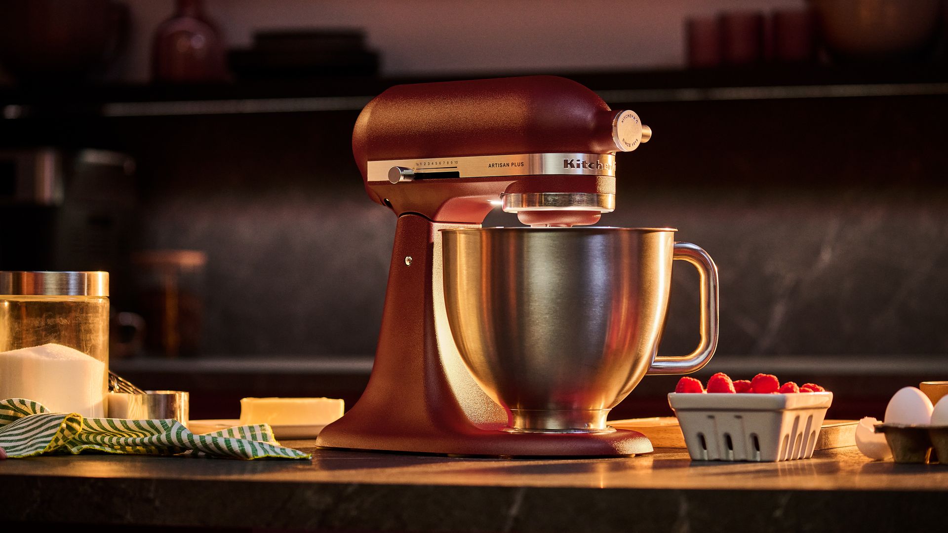 Red KitchenAid stand mixer with a stainless steel bowl on a kitchen counter; left: a sugar jar and striped towel, right: raspberries in a basket and eggs in a carton.