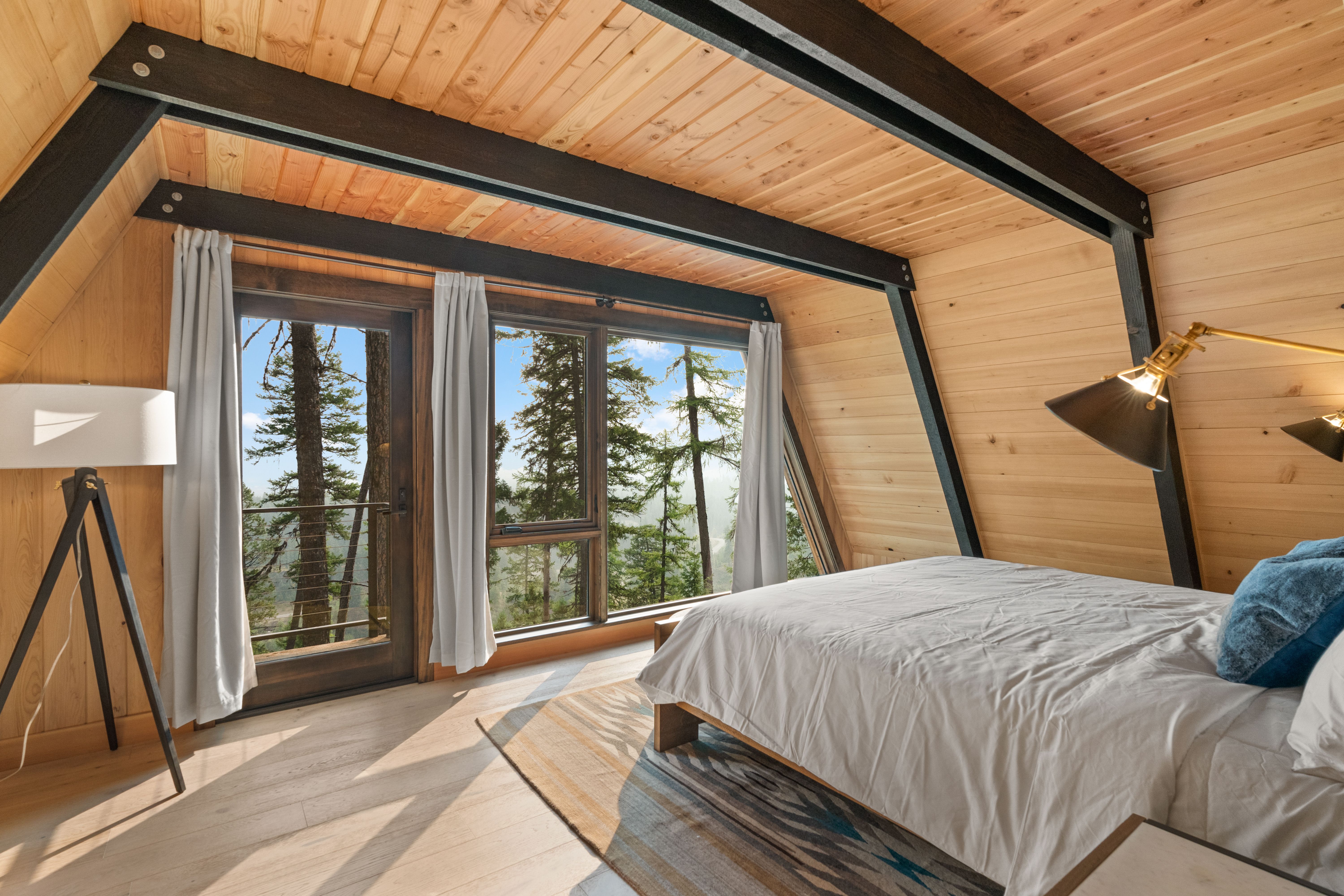 A bed sits to the right and large windows show trees outside.