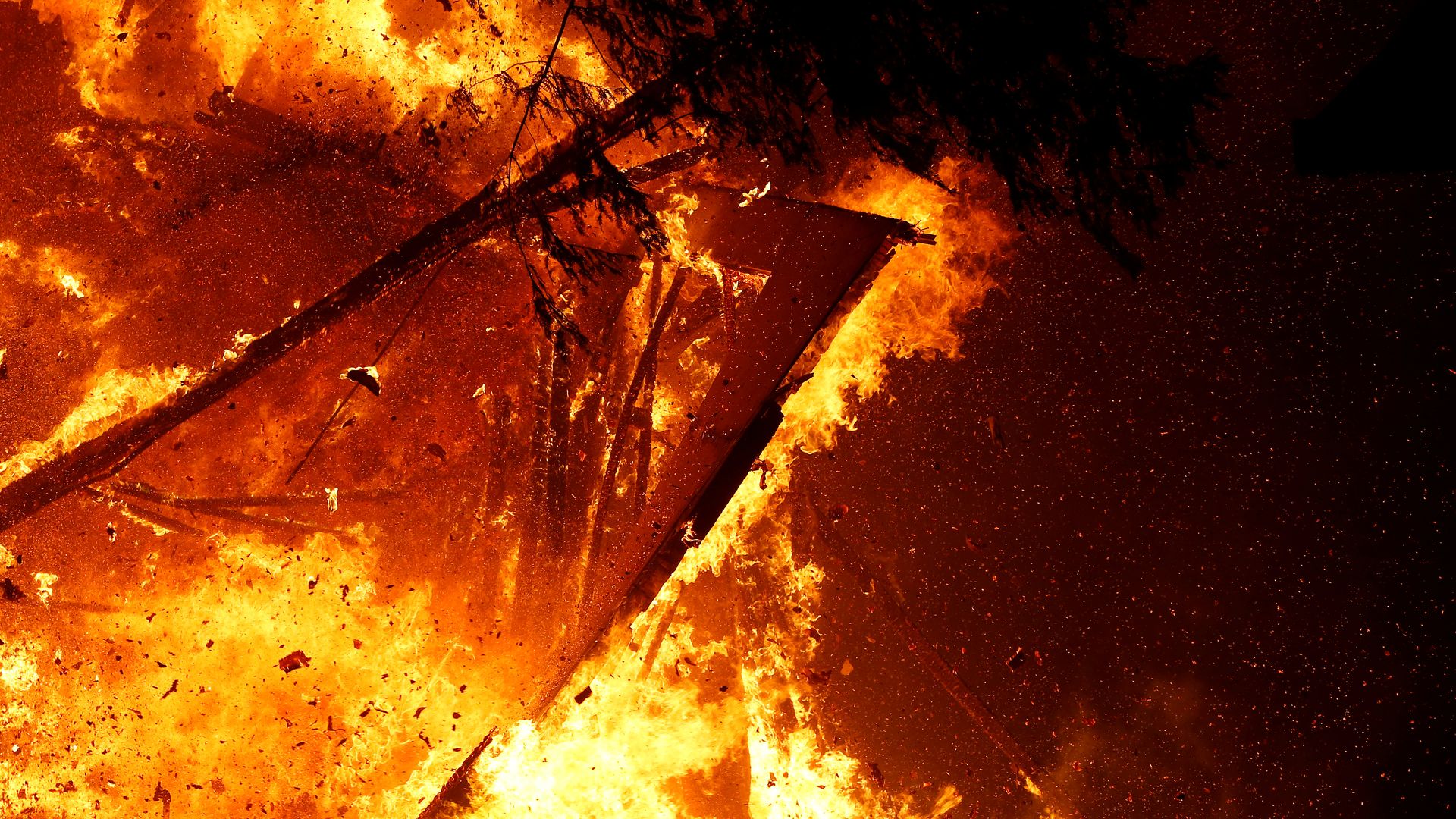 In this image, a tree and a crumbling structure, both on fire, fall to the ground in the dark