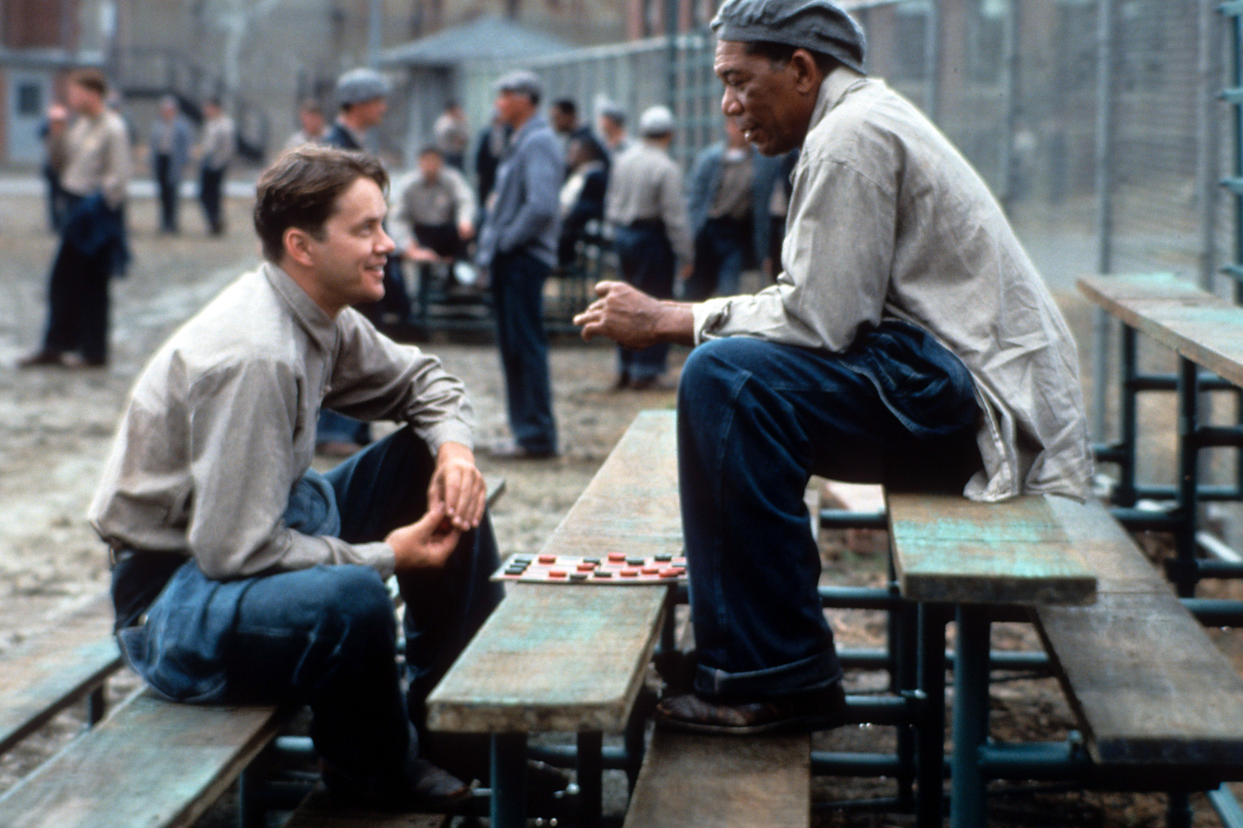 Two inmates sit on a bench in a prison yard, smiling while playing checkers, with other prisoners and a chain-link fence in the background.