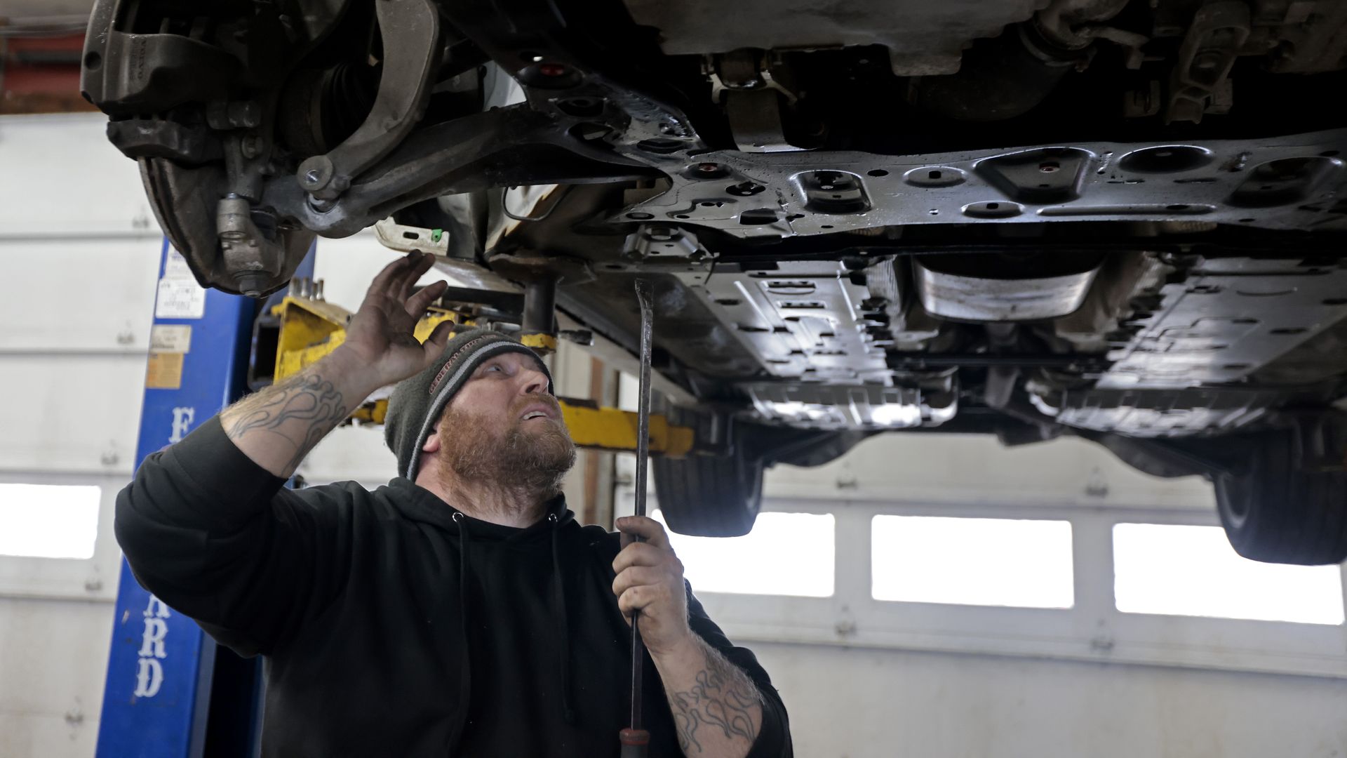 An auto mechanic looks at the undercarriage of a car while it's hoisted up