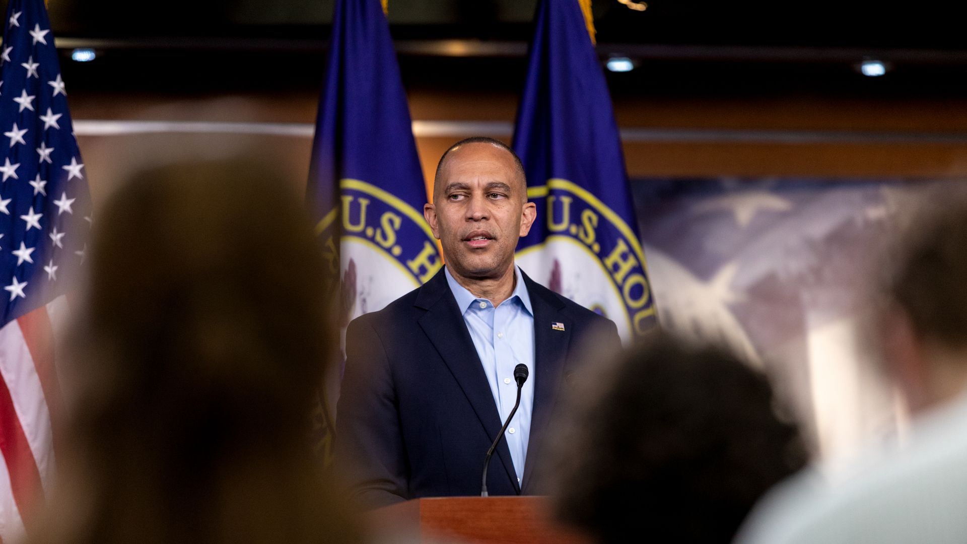 Hakeem Jeffries, wearing a tieless blue suit, speaks at a wooden podium in front of several flags and behind the blurry silhouettes of several reporters.