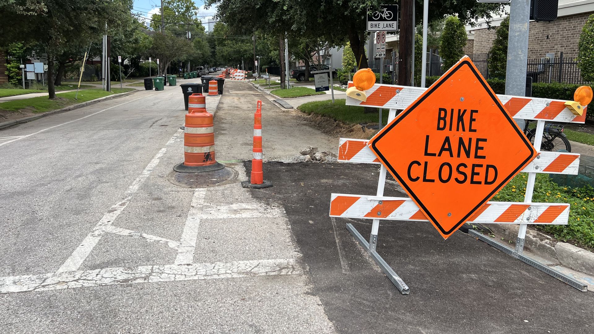Orange diamond sign reading "BIKE LANE CLOSED" in front of a construction area with orange barrels and cones on a street lined with trees and houses 
