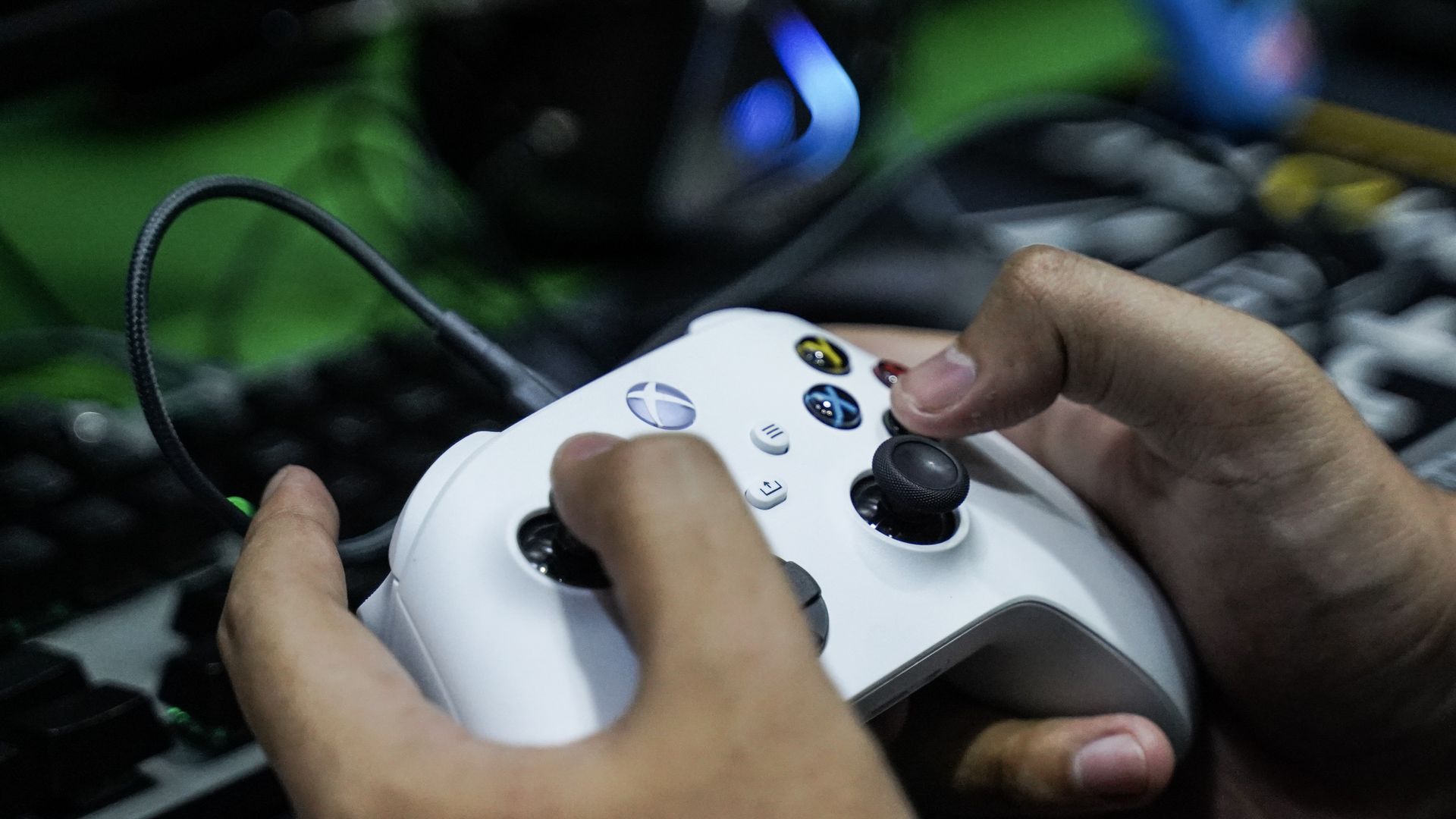 Close-up of hands gripping a white Xbox controller with joysticks, colored buttons, and a wired cable; blurred green background suggests a gaming setup.