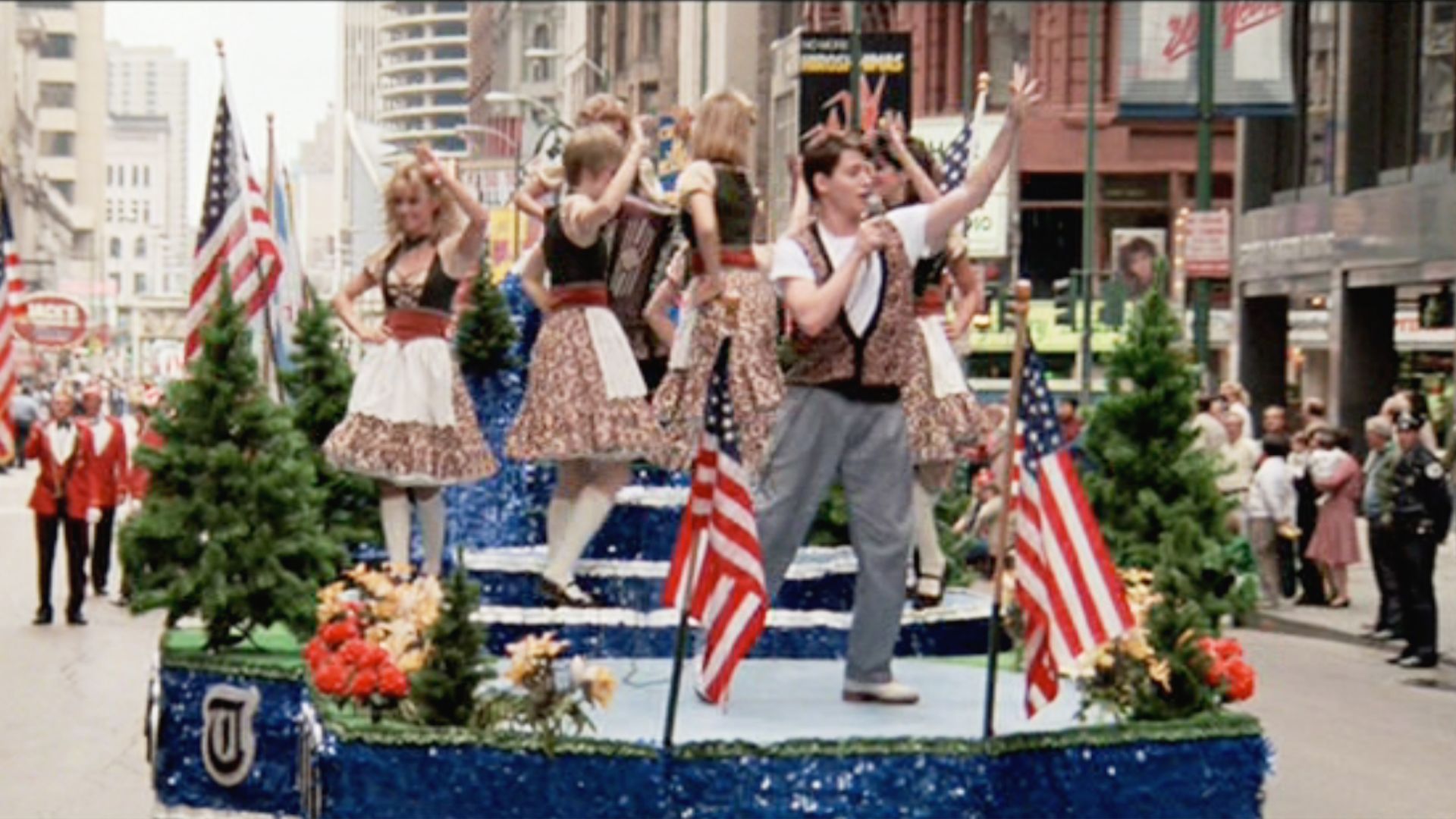 A street parade float in blue, with performers in 1950s outfits dancing and carrying American flags. A man sings into a microphone as spectators watch amid city buildings.