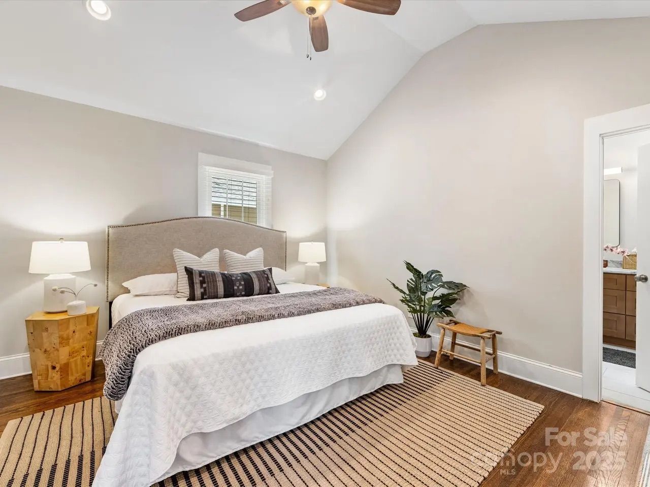 Bright bedroom with beige walls, king bed with light gray headboard, white bedding, dark gray throw, two lamps on wooden bedside tables, striped rug, ceiling fan, and a small bench with potted plant.